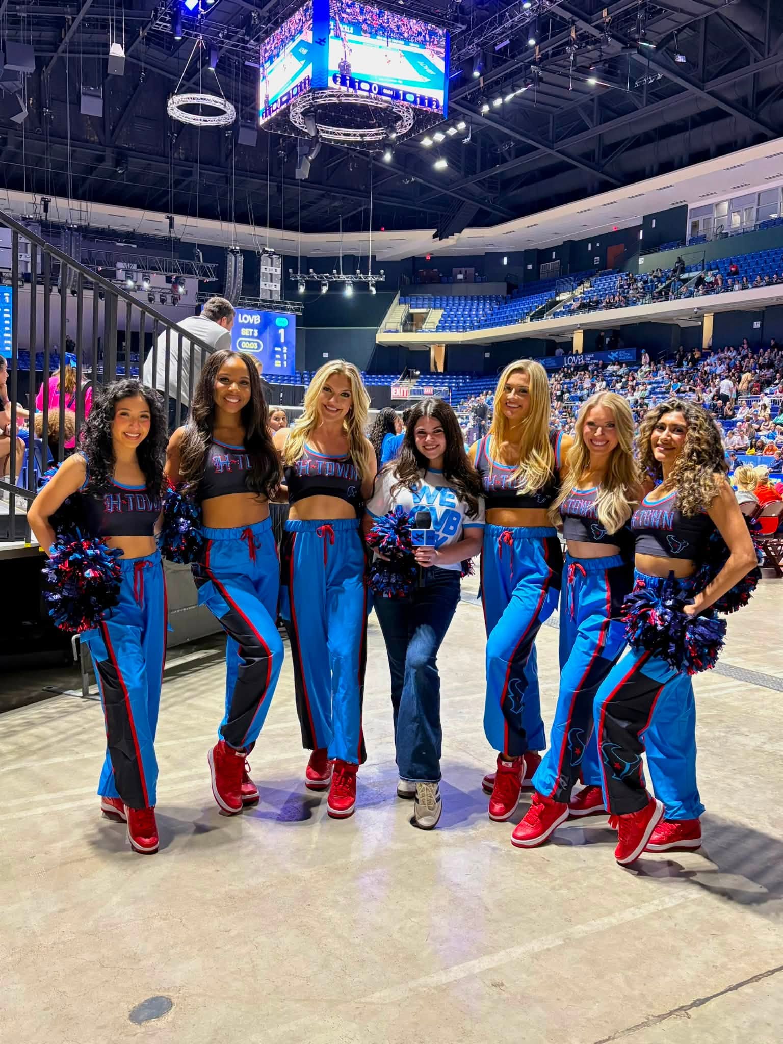 Group of seven cheerleaders in blue and black uniforms with pom-poms, standing in an indoor arena with audience and screens.