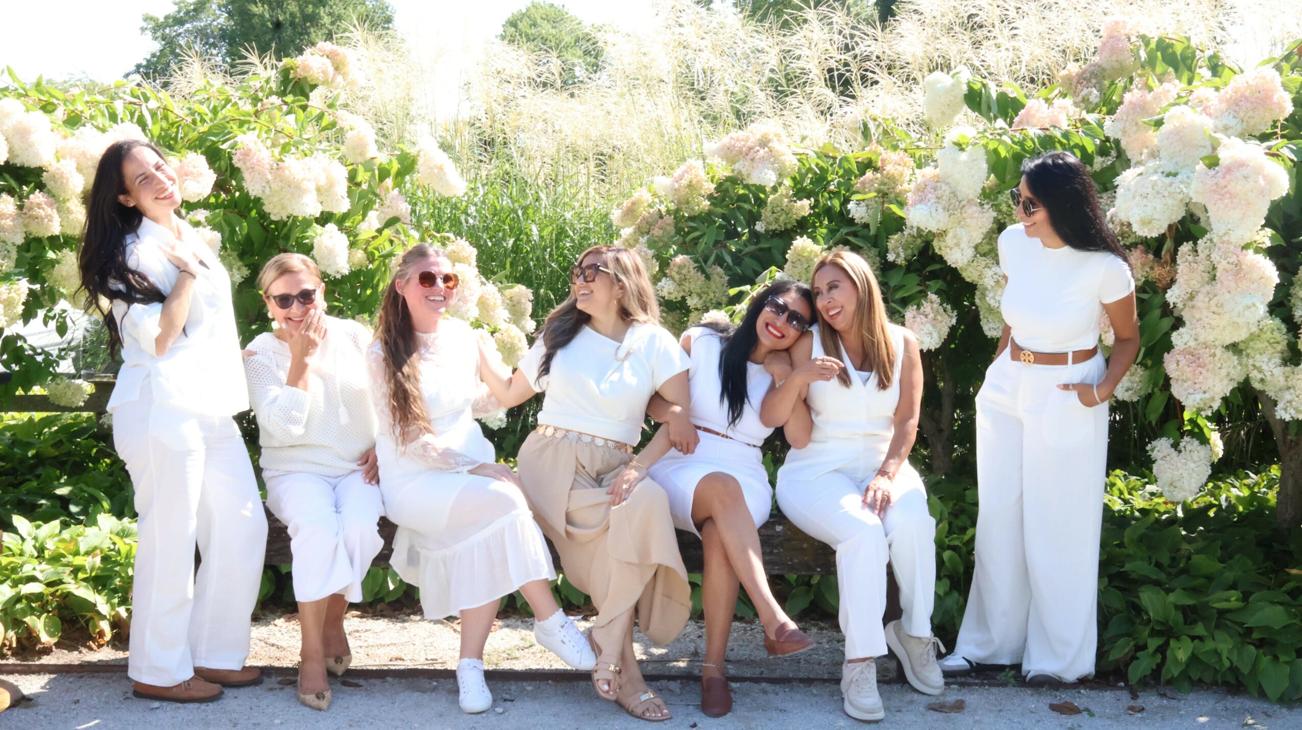 Group of women sitting and standing outdoors among white flowers, smiling and enjoying the sunny day.