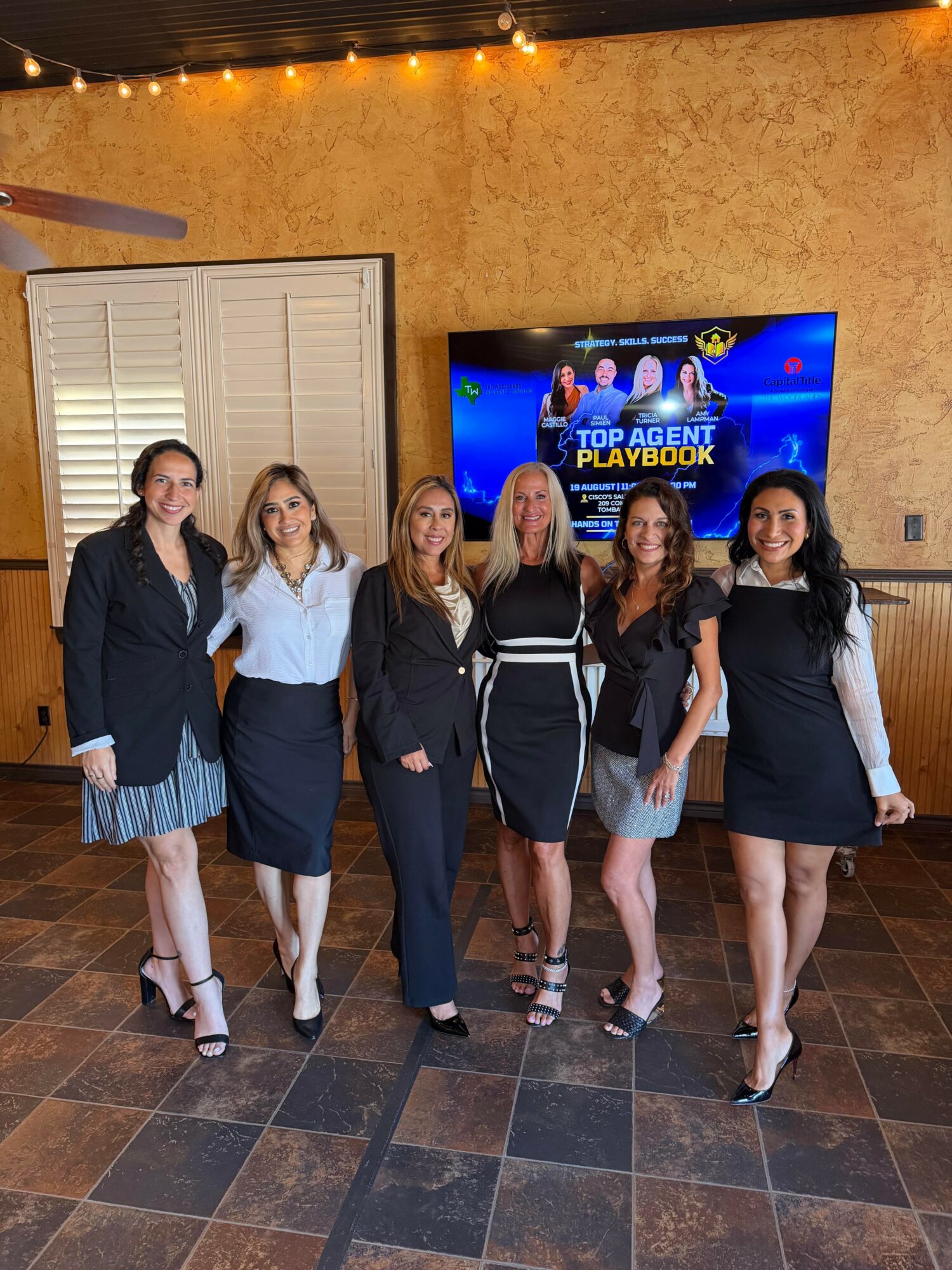 Six women stand together indoors, smiling, in front of a TV screen displaying 'Top Agent Playbook' with a blue background.