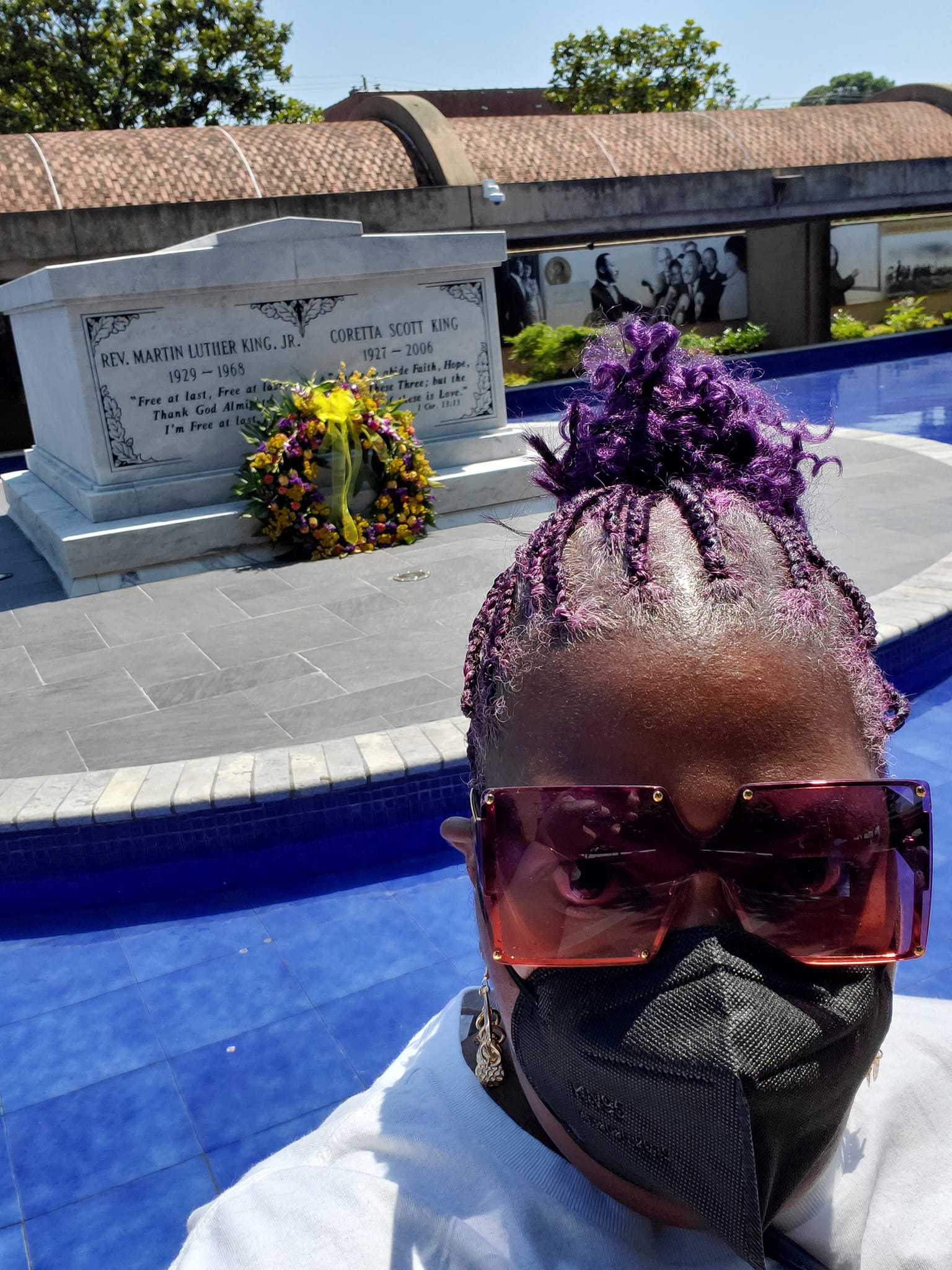 Person with purple braided hair, large sunglasses, and black face mask in front of a memorial with flowers.