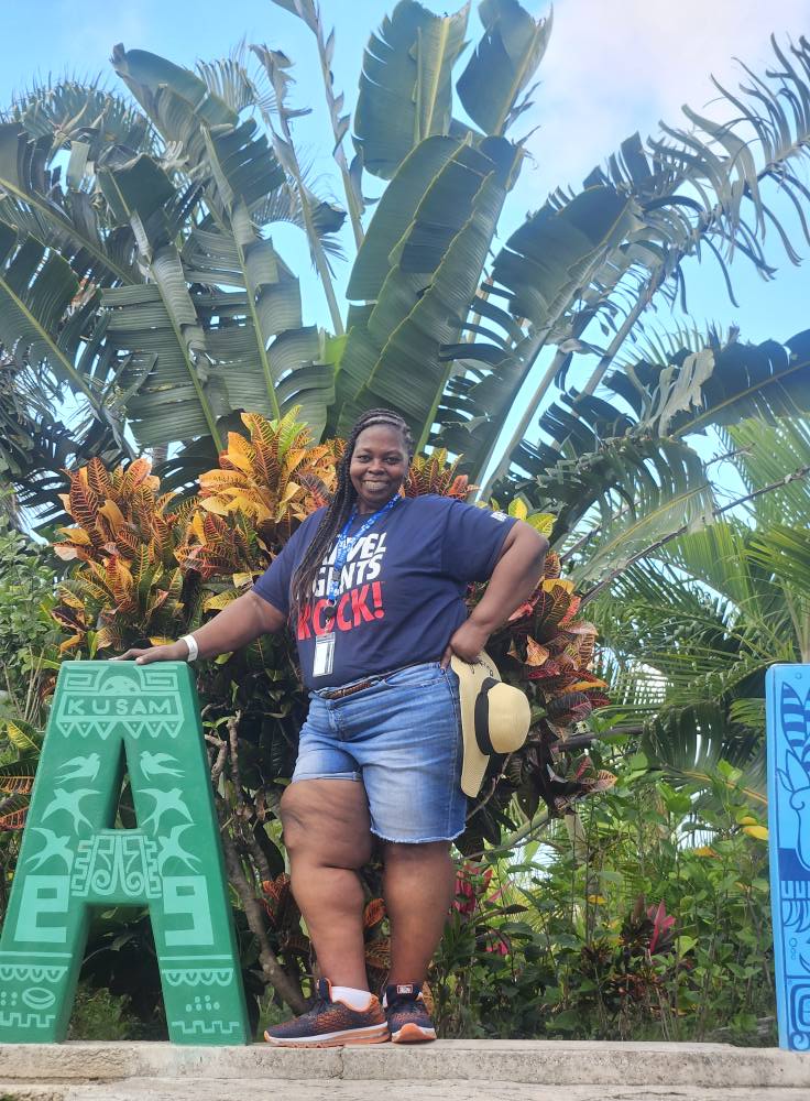 Person standing outdoors in front of large tropical plants, smiling, wearing a dark T-shirt and denim shorts, holding a hat.