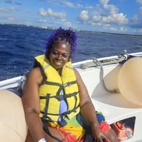 Woman with purple hair wearing a yellow life jacket on a boat, smiling, with water and sky in background.