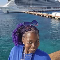Woman with purple hair and sunglasses smiling near a dock with a large cruise ship in the background.