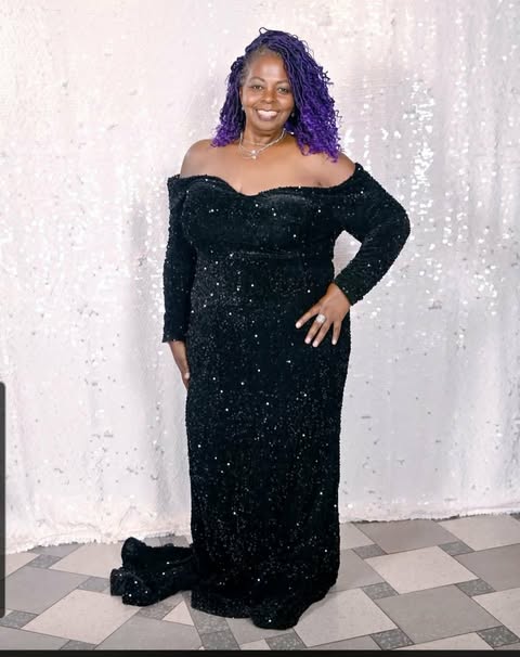 Woman in a black off-shoulder dress with purple curly hair, smiling, standing against a white textured background.