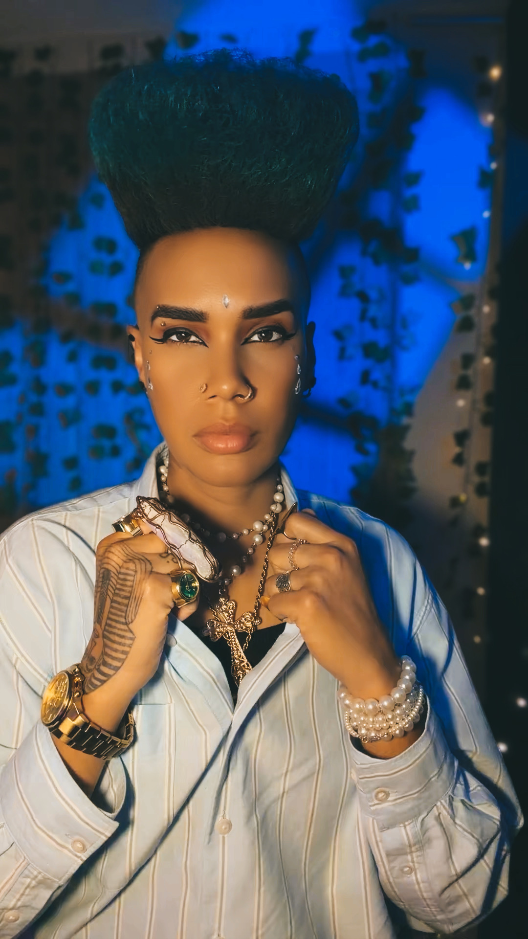 Woman with tall hairstyle, wearing jewelry and a striped shirt, looking at the camera against a blue background.
