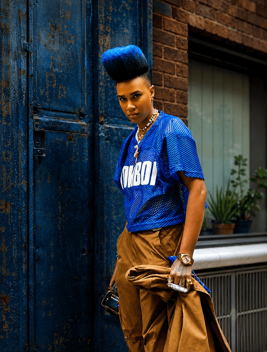 Woman with tall blue hairstyle wearing a blue sports jersey and brown pants, standing outdoors near a blue door and brick wall.