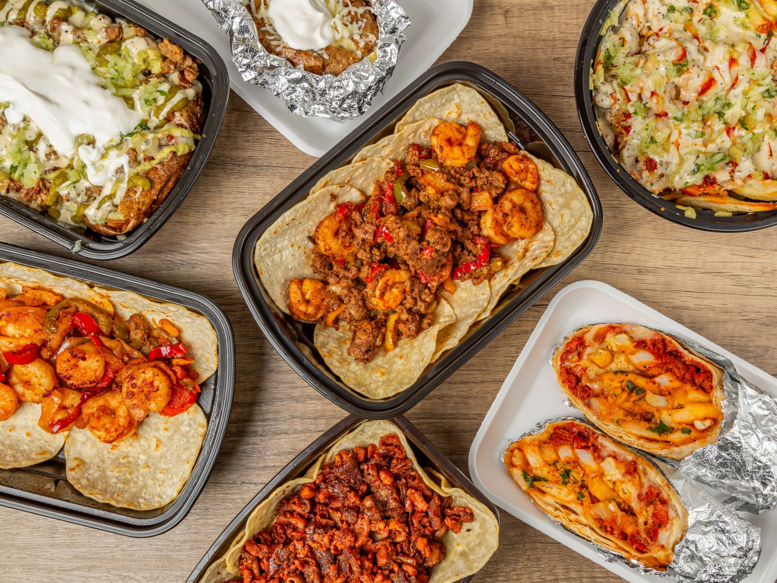 Multiple takeout containers with various dishes, including tacos, rice, and meat, on a wooden surface.