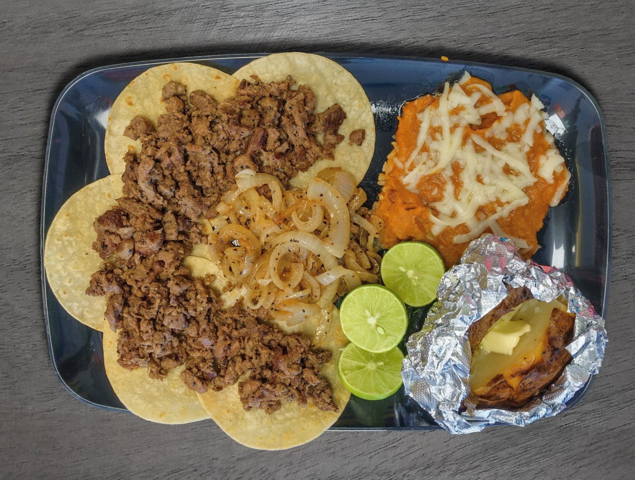 Tray with ground beef, onions, shredded cheese, two lime slices, and a wrapped tamale on a dark surface.
