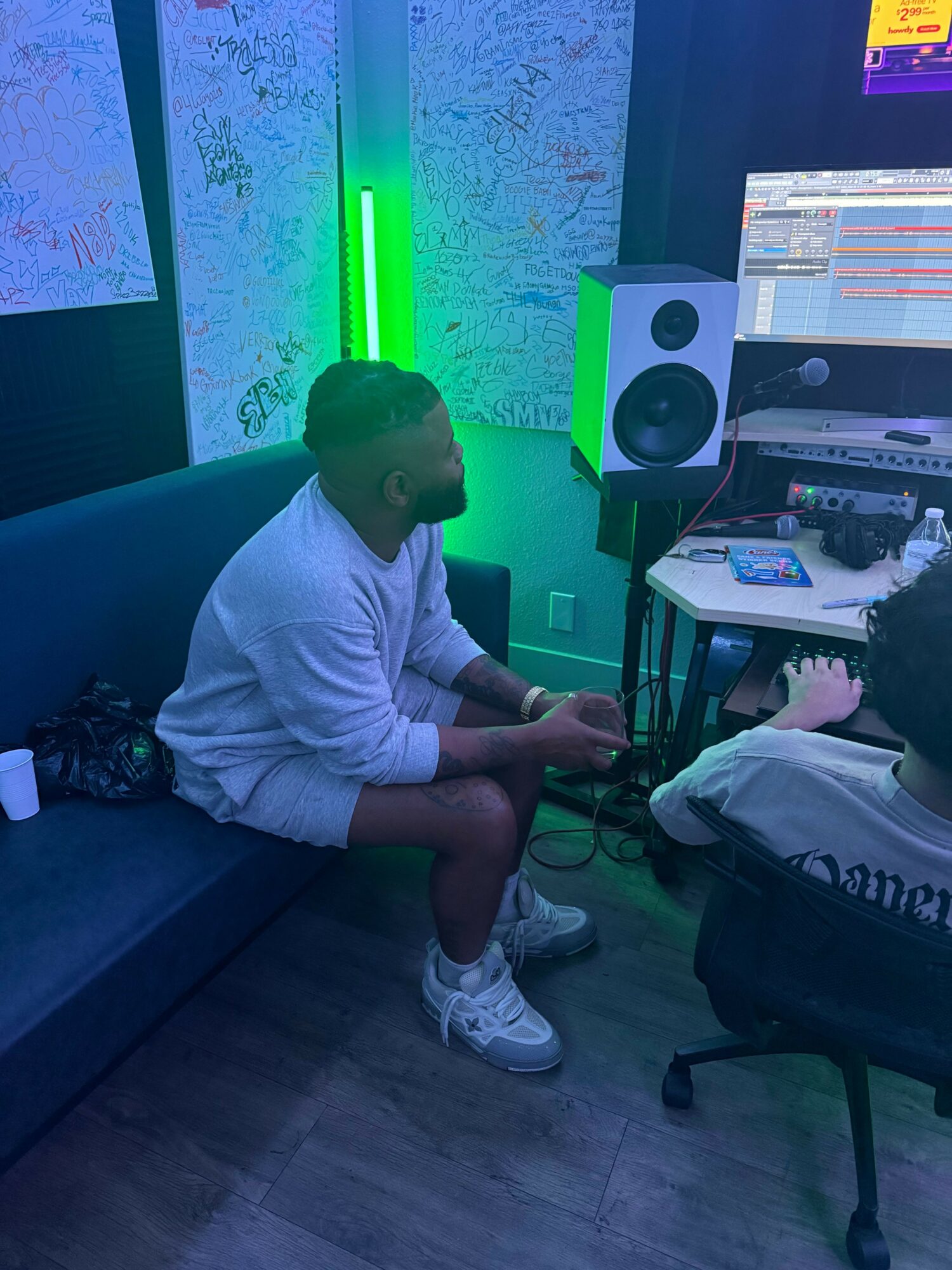 Person sitting on a blue couch in a music studio with green lighting, working at a computer with speakers and equipment.