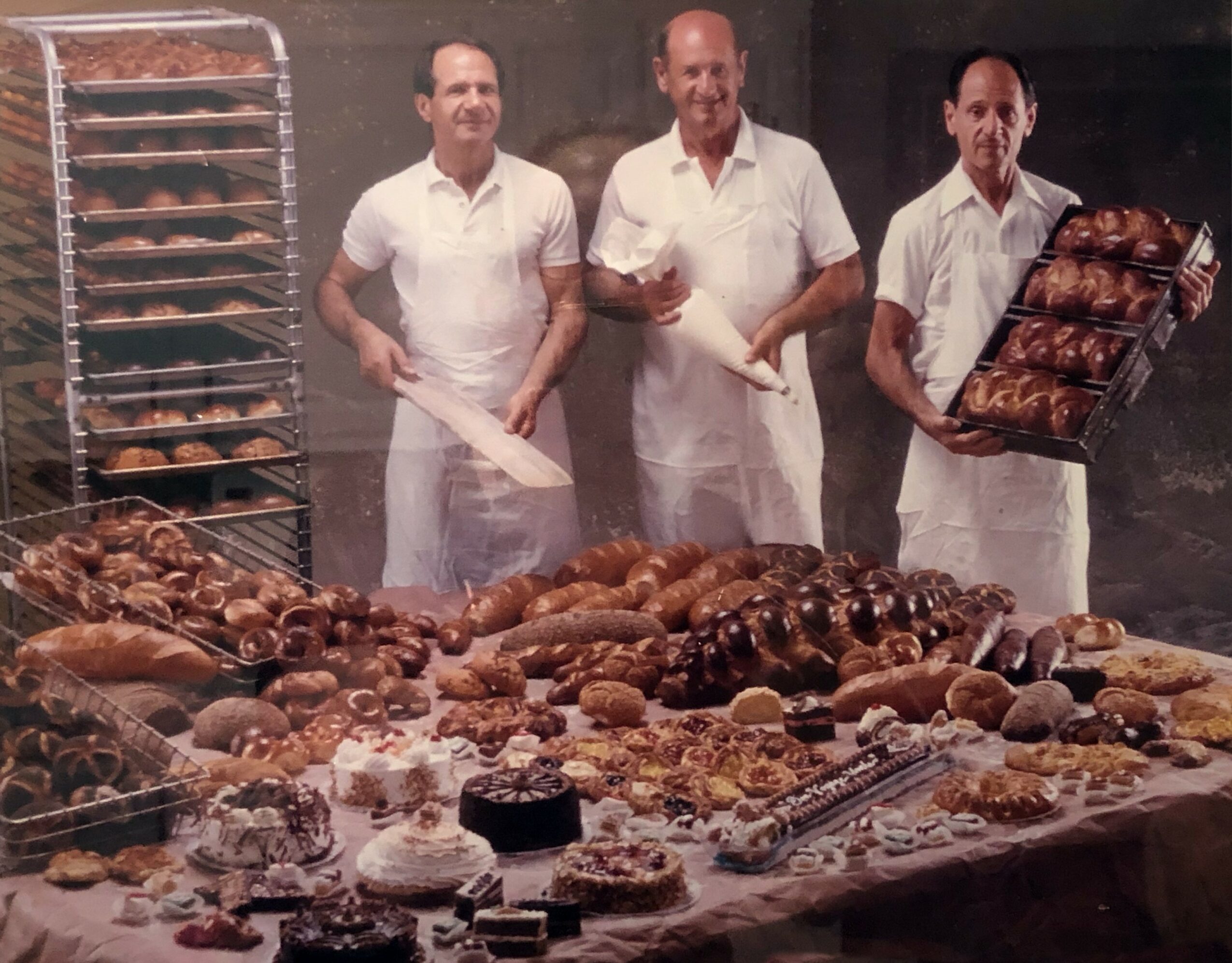 Three bakers stand behind a large table filled with baked goods, holding trays and tools, in a bakery setting.
