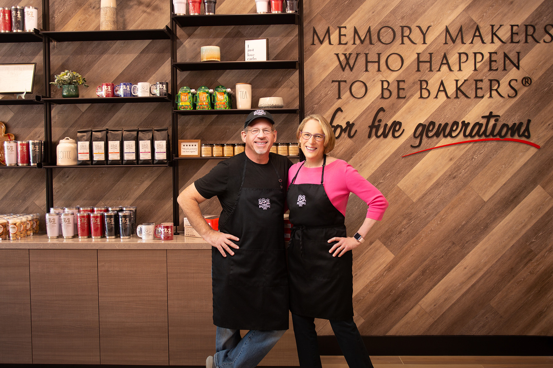 Two people stand in front of shelves with baking supplies, smiling, with a wall that has text about memory makers and bakers.