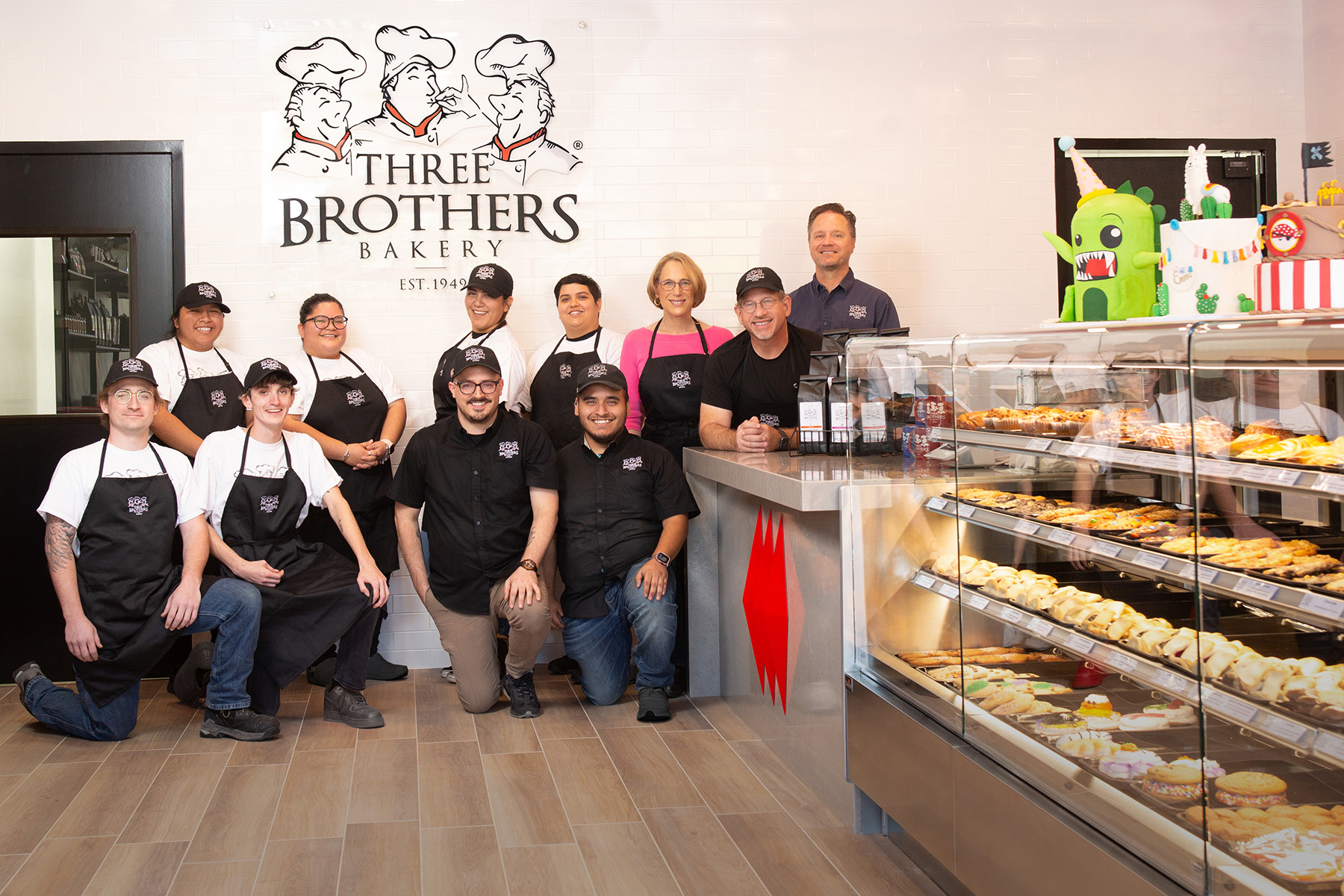 Group of bakery staff and customers inside Three Brothers Bakery, with display case of baked goods and bakery sign in background.
