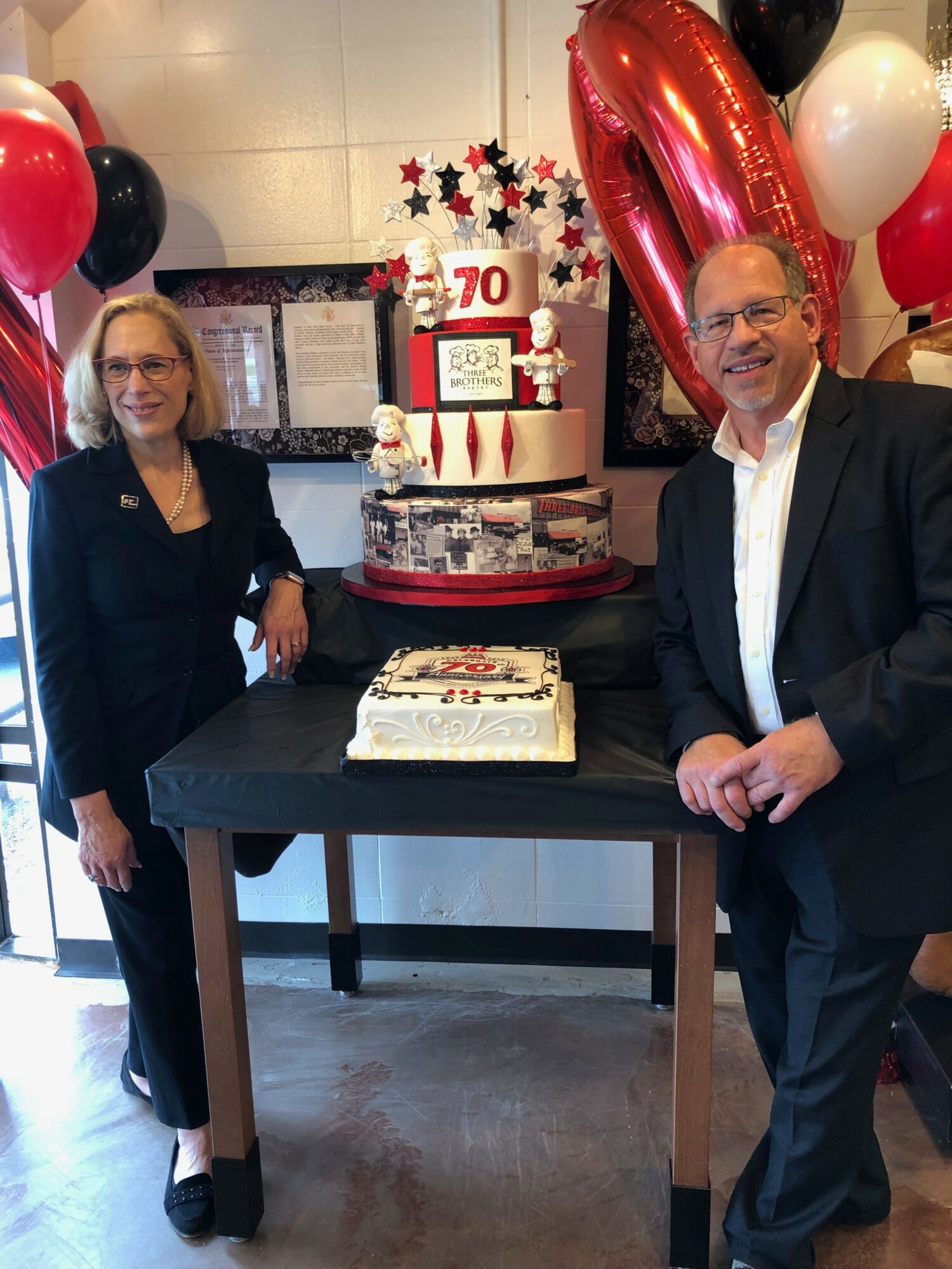 Two people stand beside a table with two cakes, surrounded by red, black, and white balloons, celebrating a 70th birthday.