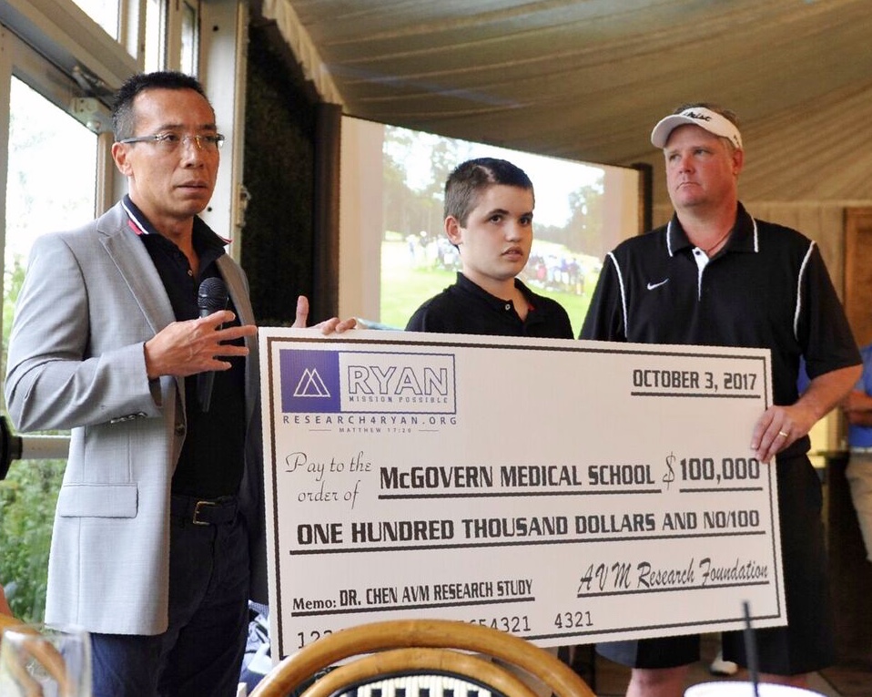 Three men holding a large check, with a man speaking into a microphone, in an indoor setting with windows and a ceiling. Alt text word count: 44, confidence: 0.95