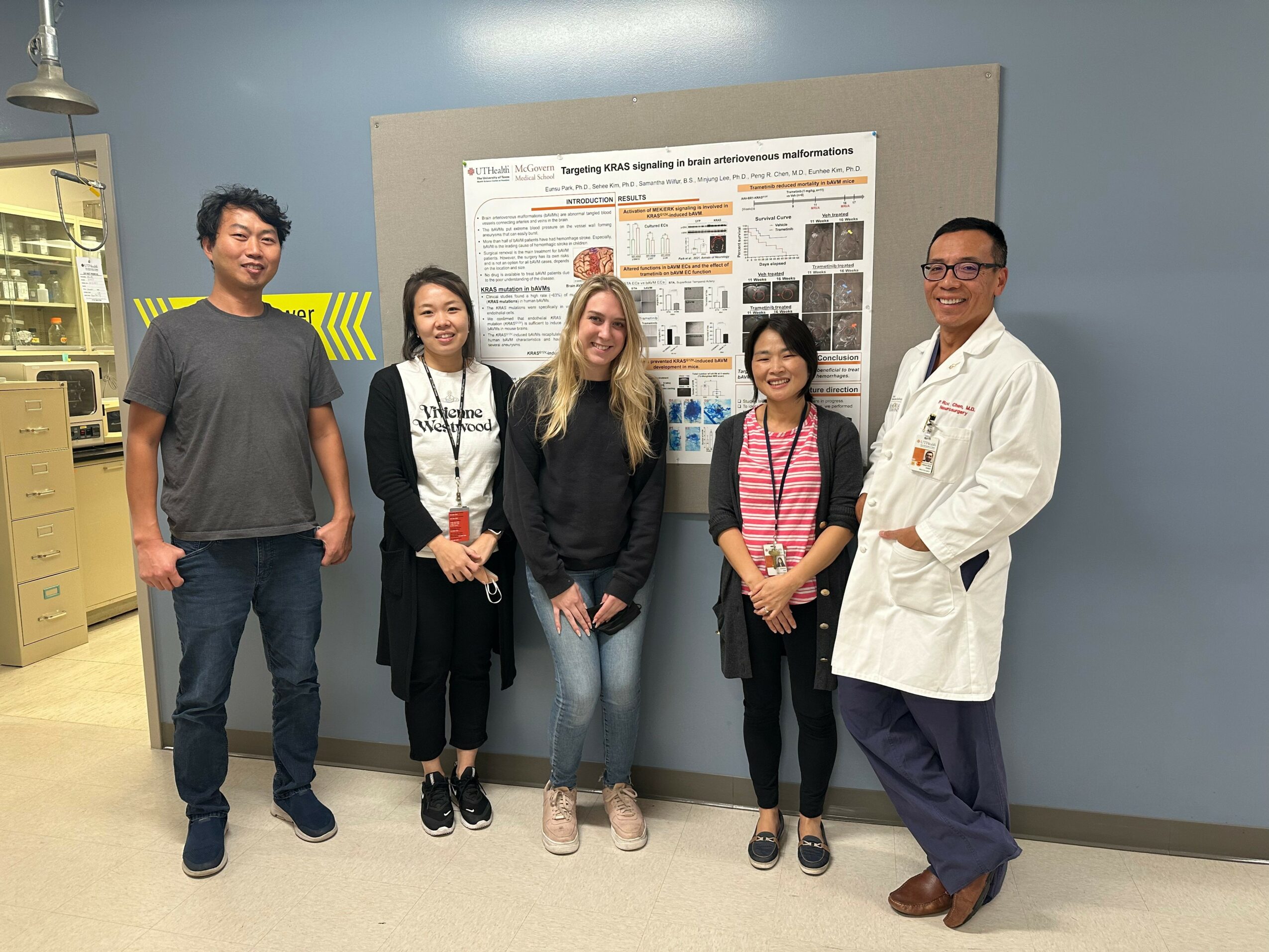 Five people standing in front of a research poster, smiling, in a room with blue walls and a yellow shelf.