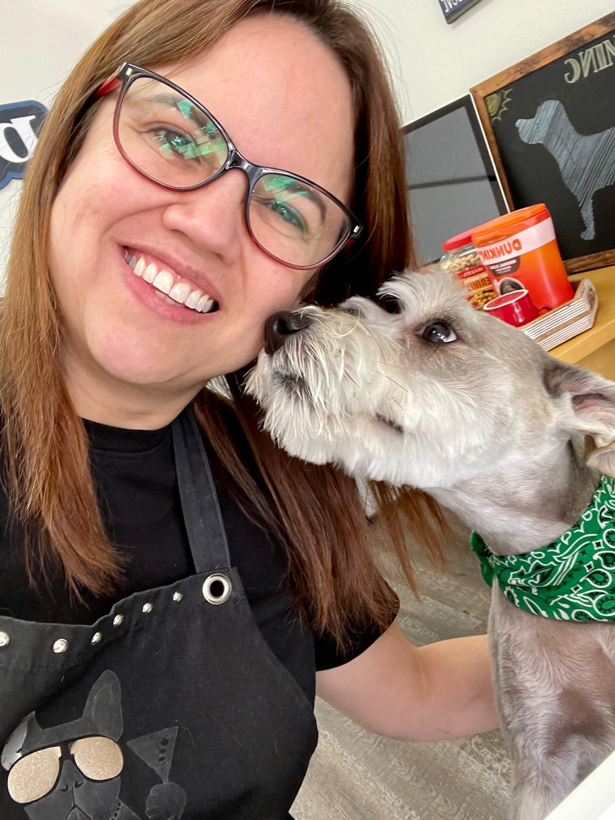 Woman with glasses smiling at a dog wearing a green bandana, indoors with shelves in background.