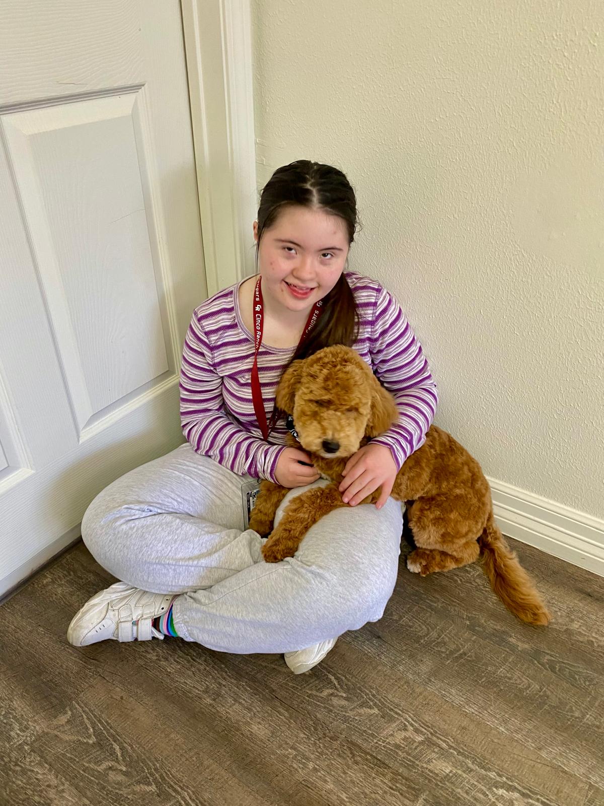 Girl sitting on floor with a brown puppy, smiling, in a room with white walls and wooden floor.