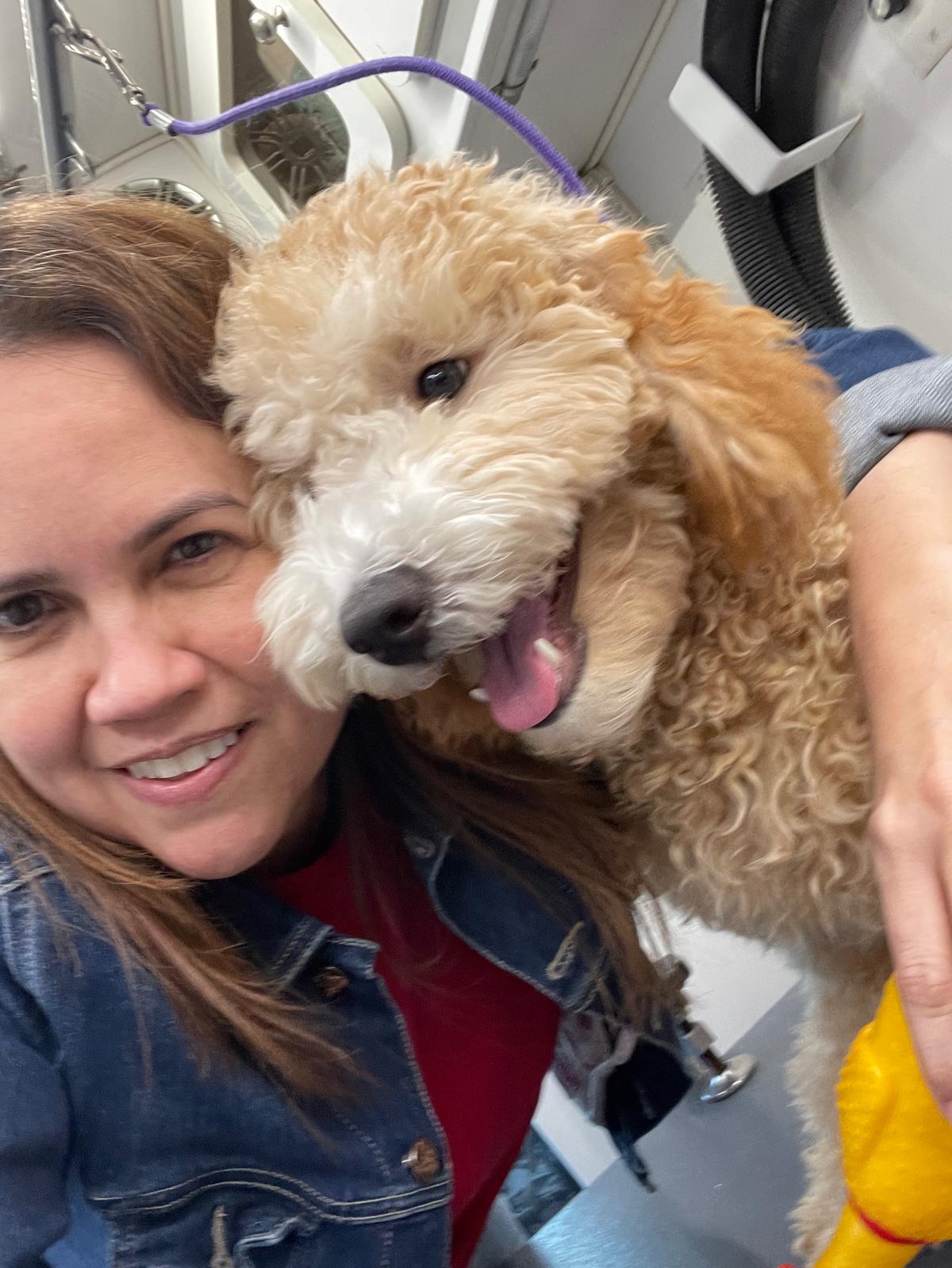 Woman smiling with a curly-haired puppy, both close-up, in a room with medical or laundry equipment visible.