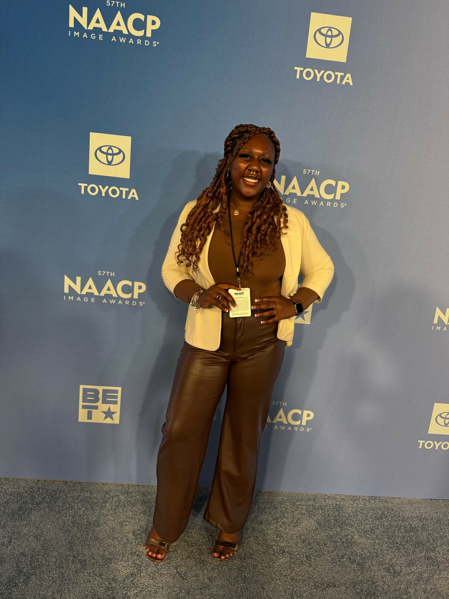 Woman with long braids smiling, standing in front of blue NAACP awards backdrop, wearing a beige blazer and brown pants.