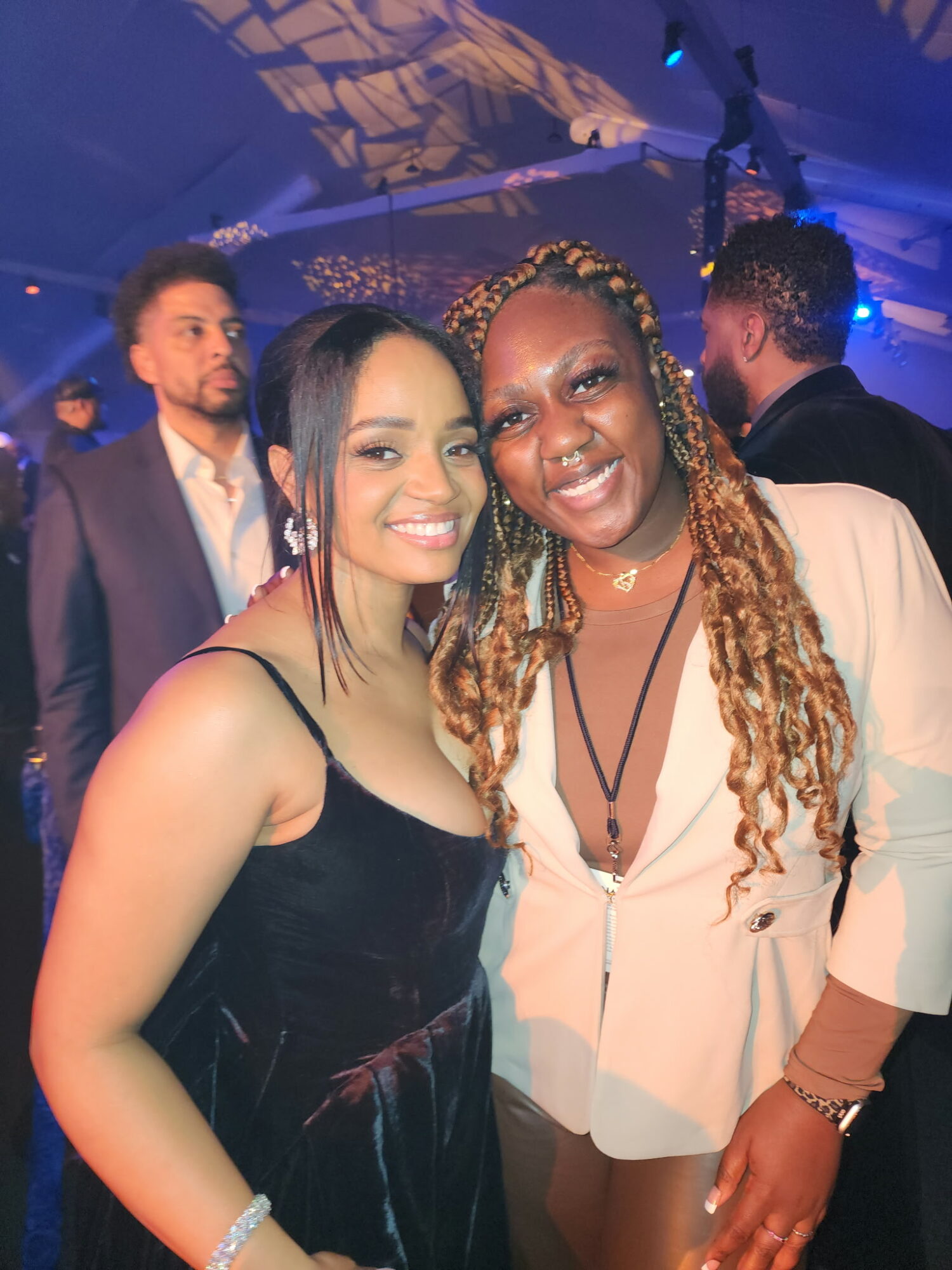 Two women smiling at a social event, with other people in the background, in a dimly lit setting.
