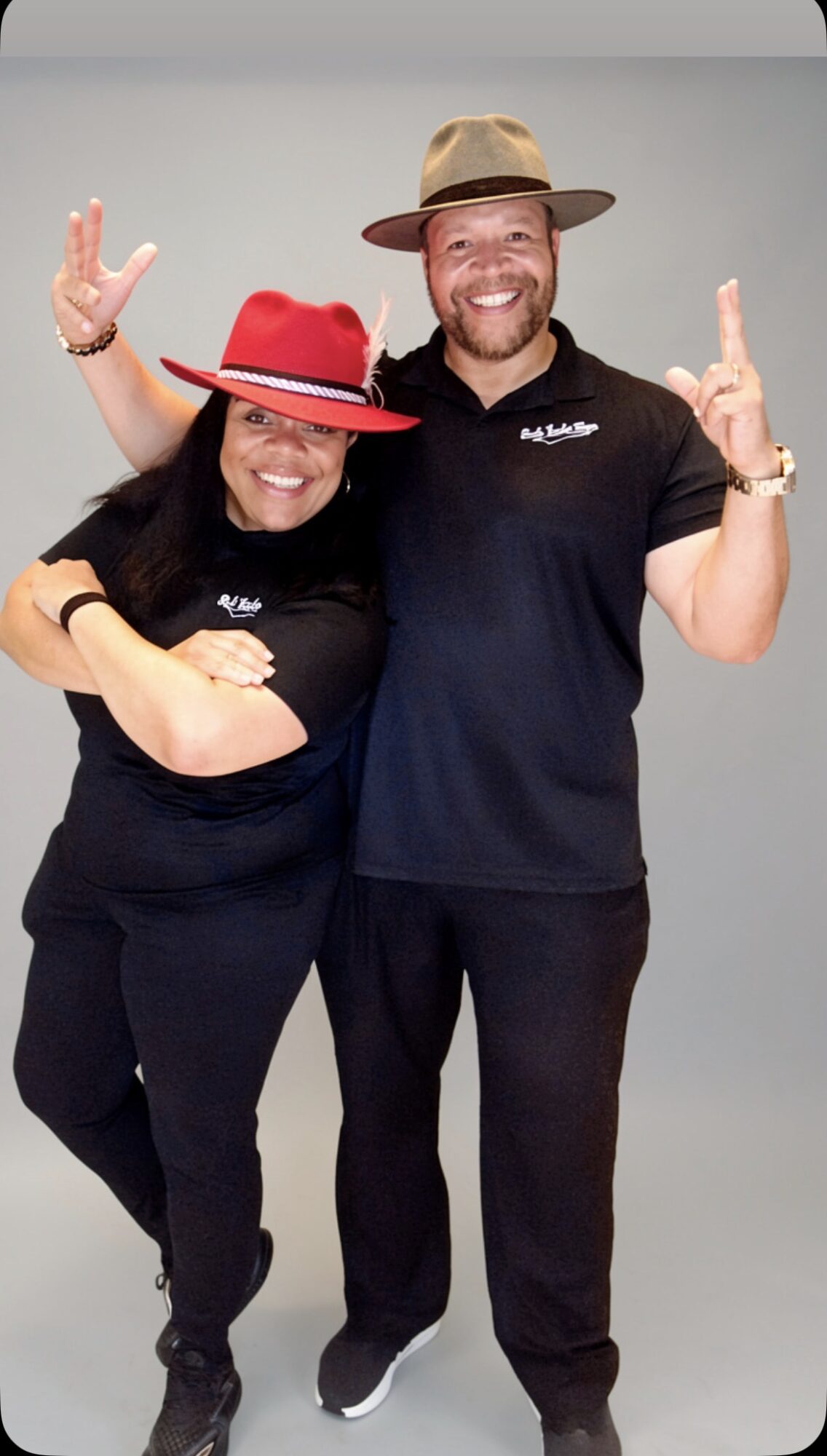 Two people smiling, wearing hats, making peace signs, standing close together against a plain background.