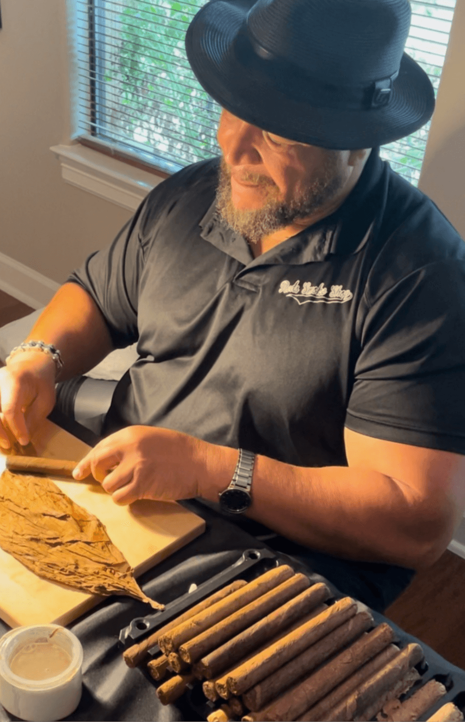 Man with a beard wearing a black hat and black polo shirt, sitting at a table with cigars and a cutting board, near a window.
