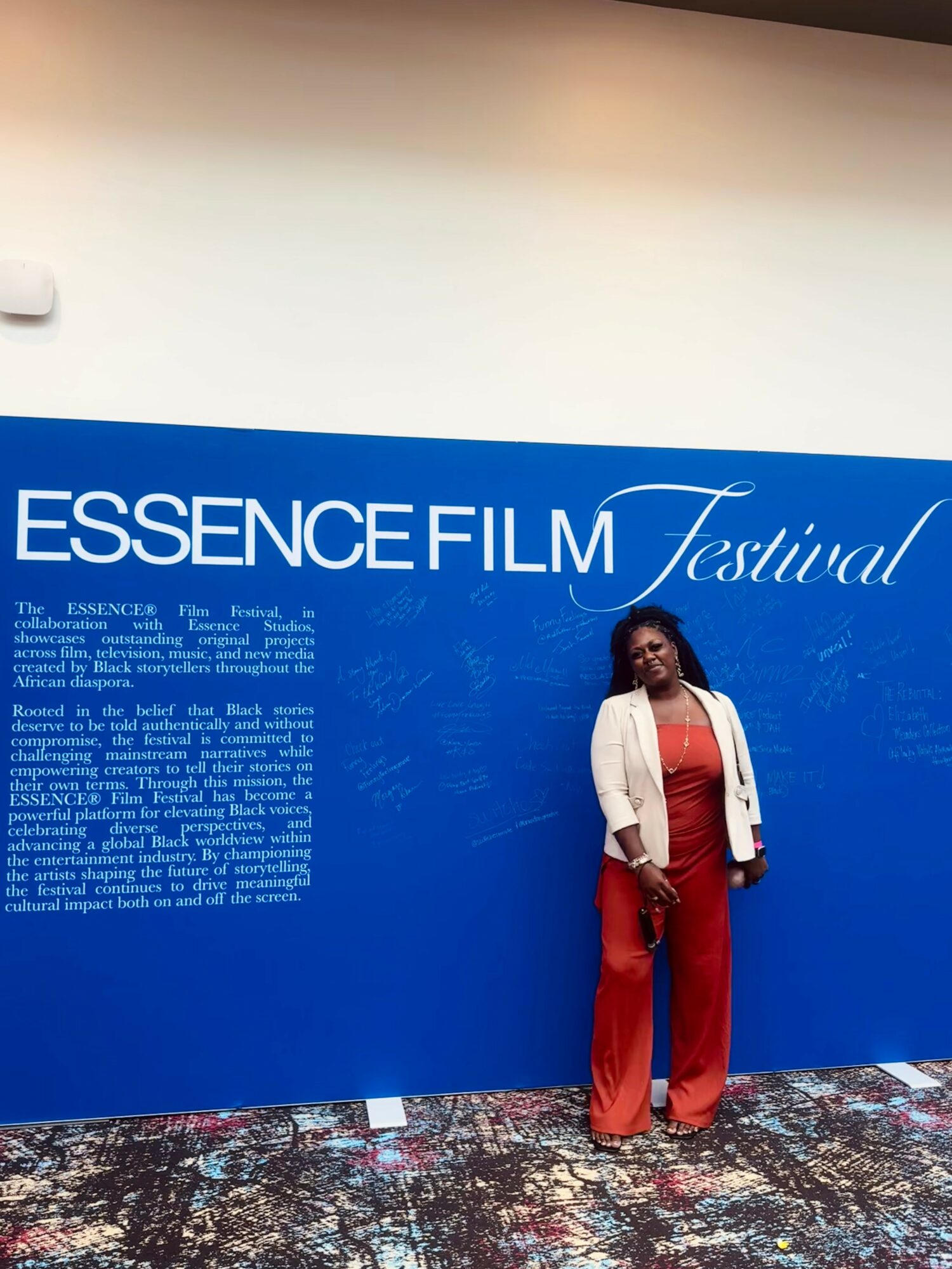 Woman standing in front of a blue wall with white text at the Essence Film Festival, wearing a white jacket and red pants.
