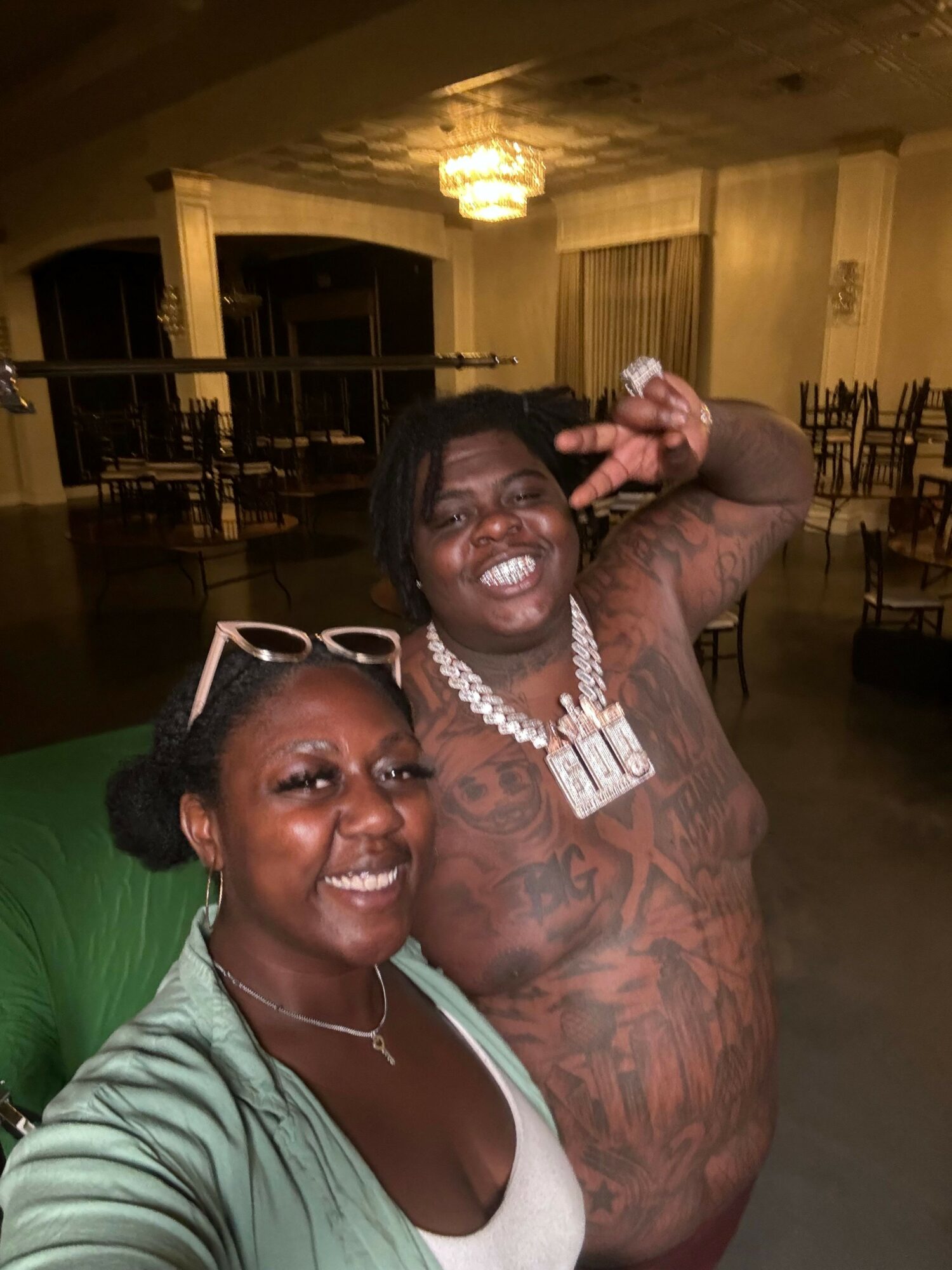 Two people taking a selfie indoors, one shirtless with tattoos and jewelry, the other wearing a light green top and sunglasses.