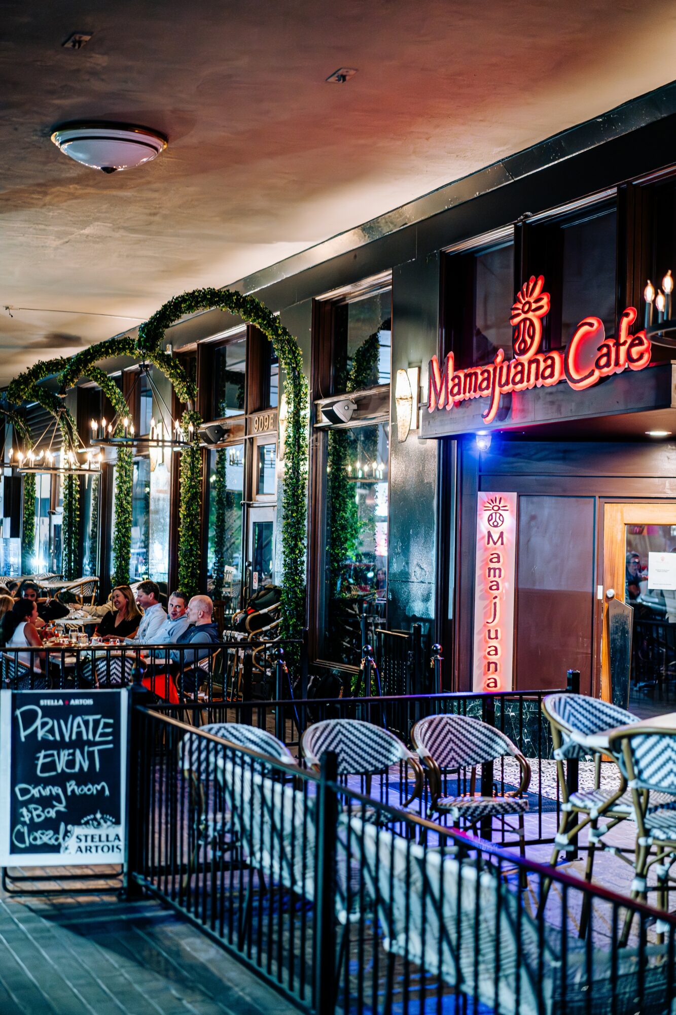 Outdoor seating area of Mamajama Cafe with tables, chairs, and a sign, under a ceiling with warm lighting.