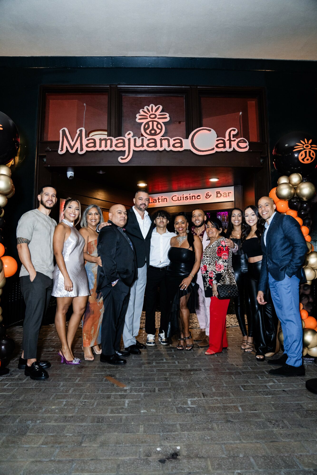 Group of people dressed in formal attire standing outside Mamajuana Cafe, with balloons and a sign above the entrance.