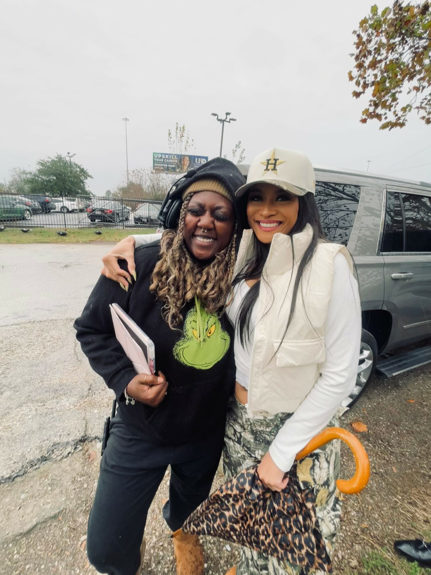 Two women smiling outdoors, one holding a notebook, with a vehicle and trees in the background.