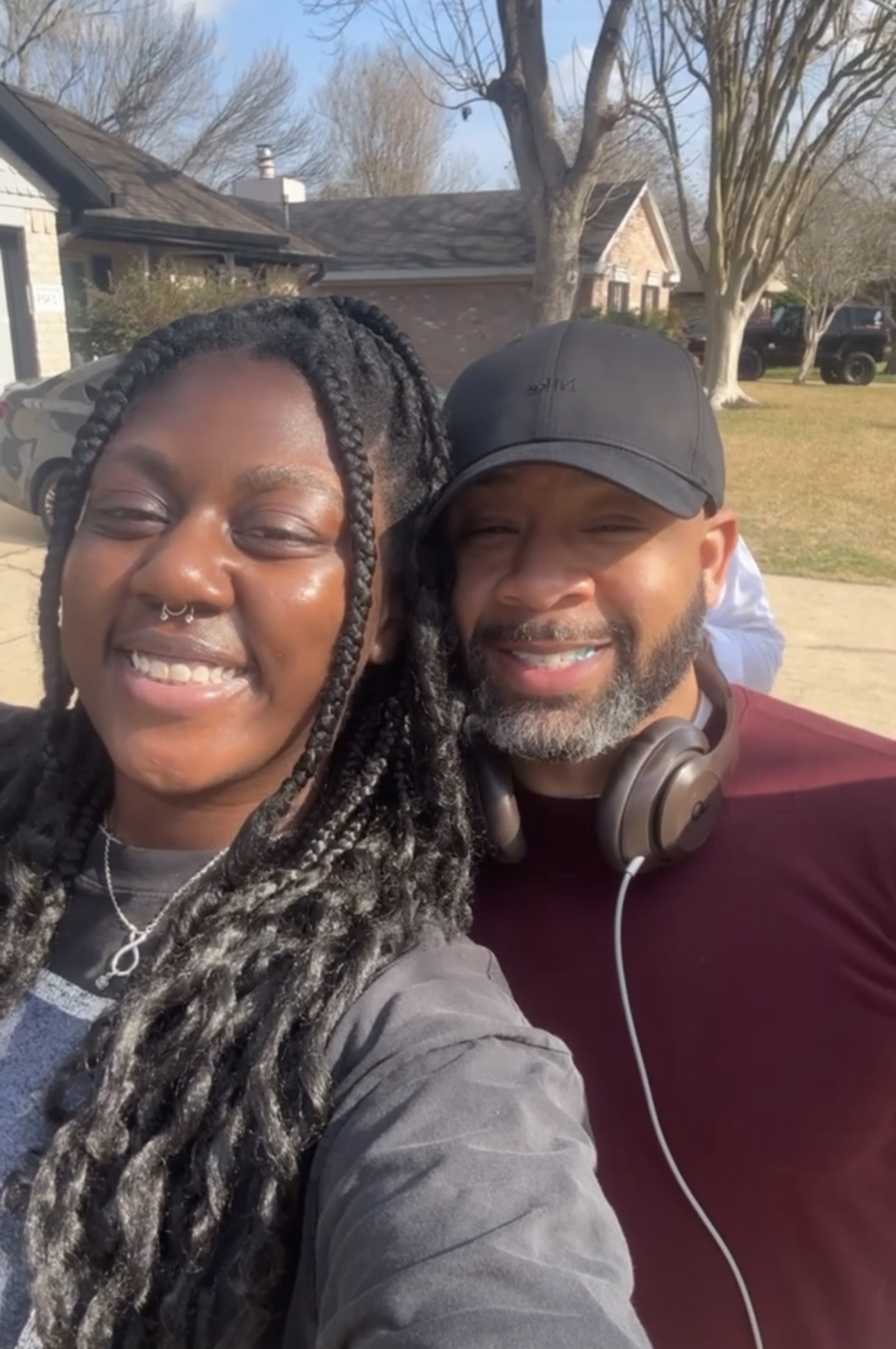Two people smiling outdoors on a sunny day, with trees and houses in the background.