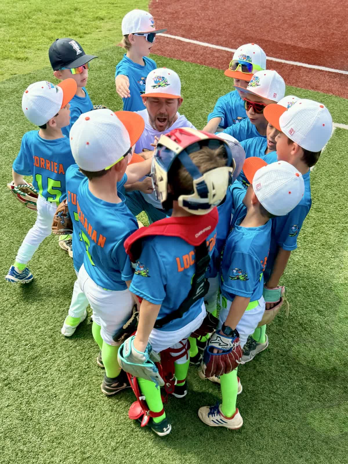 Youth baseball team in blue uniforms and white caps gathered around coach on field, coach giving instructions, players listening attentively.