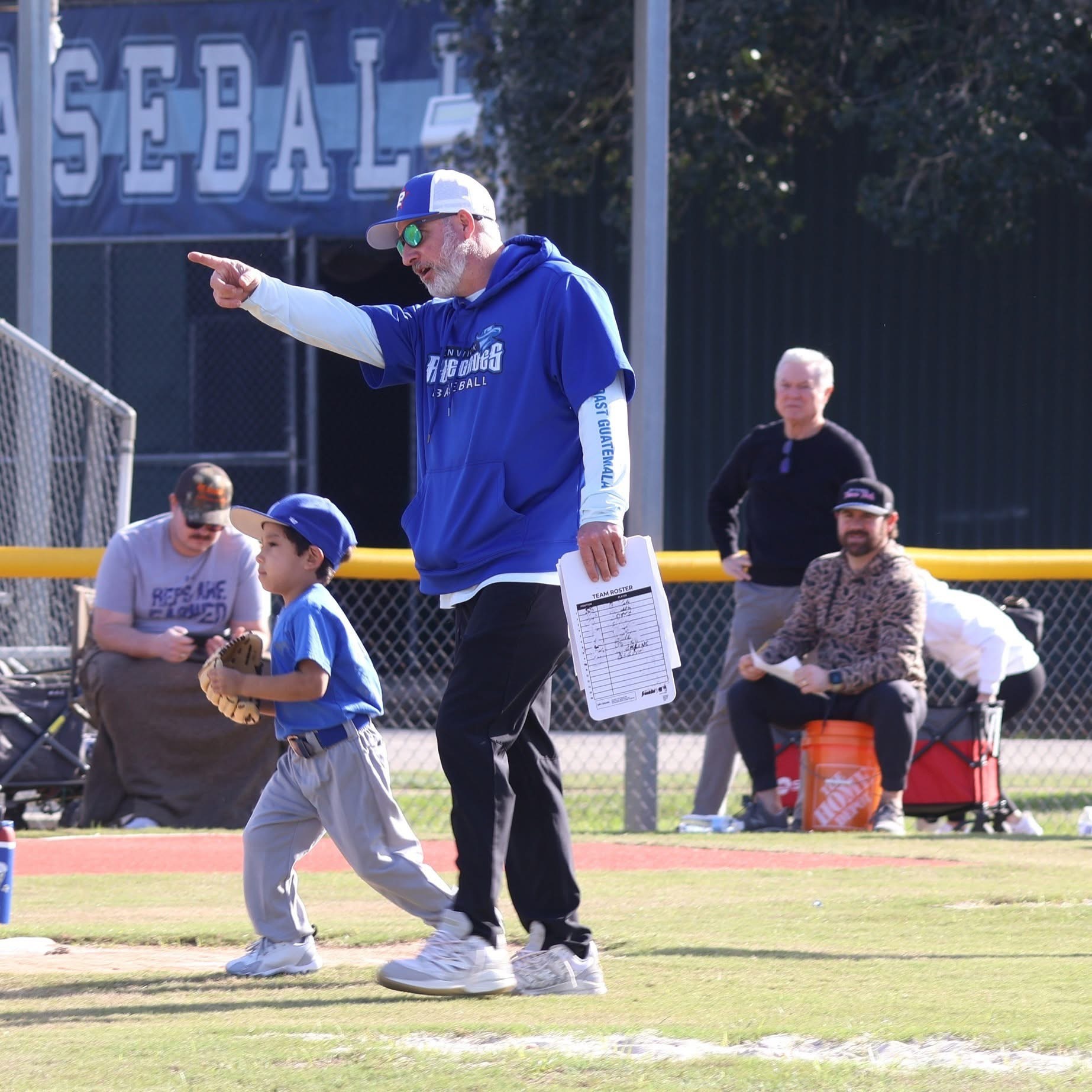 Adult coach with clipboard pointing, young player in baseball uniform on field, spectators seated behind fence, sunny day.