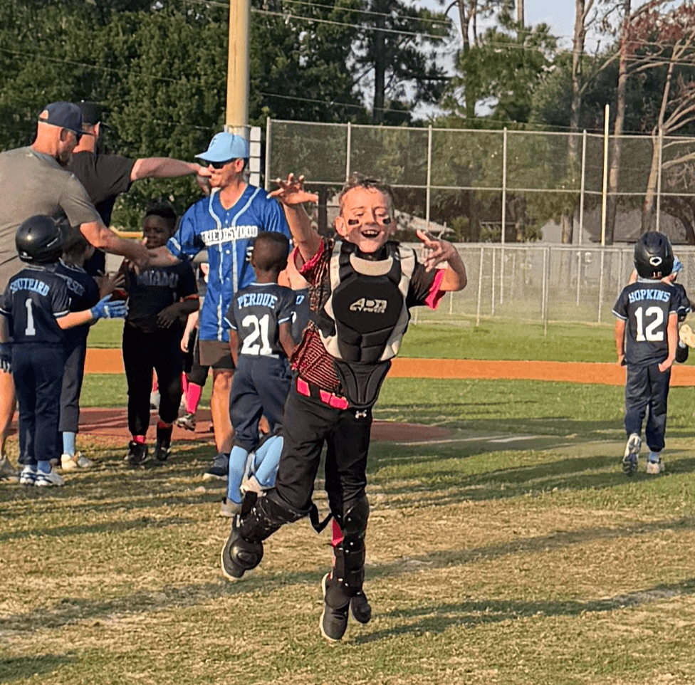 Child running towards camera with arms outstretched, smiling, on a baseball field with kids and adults in background.