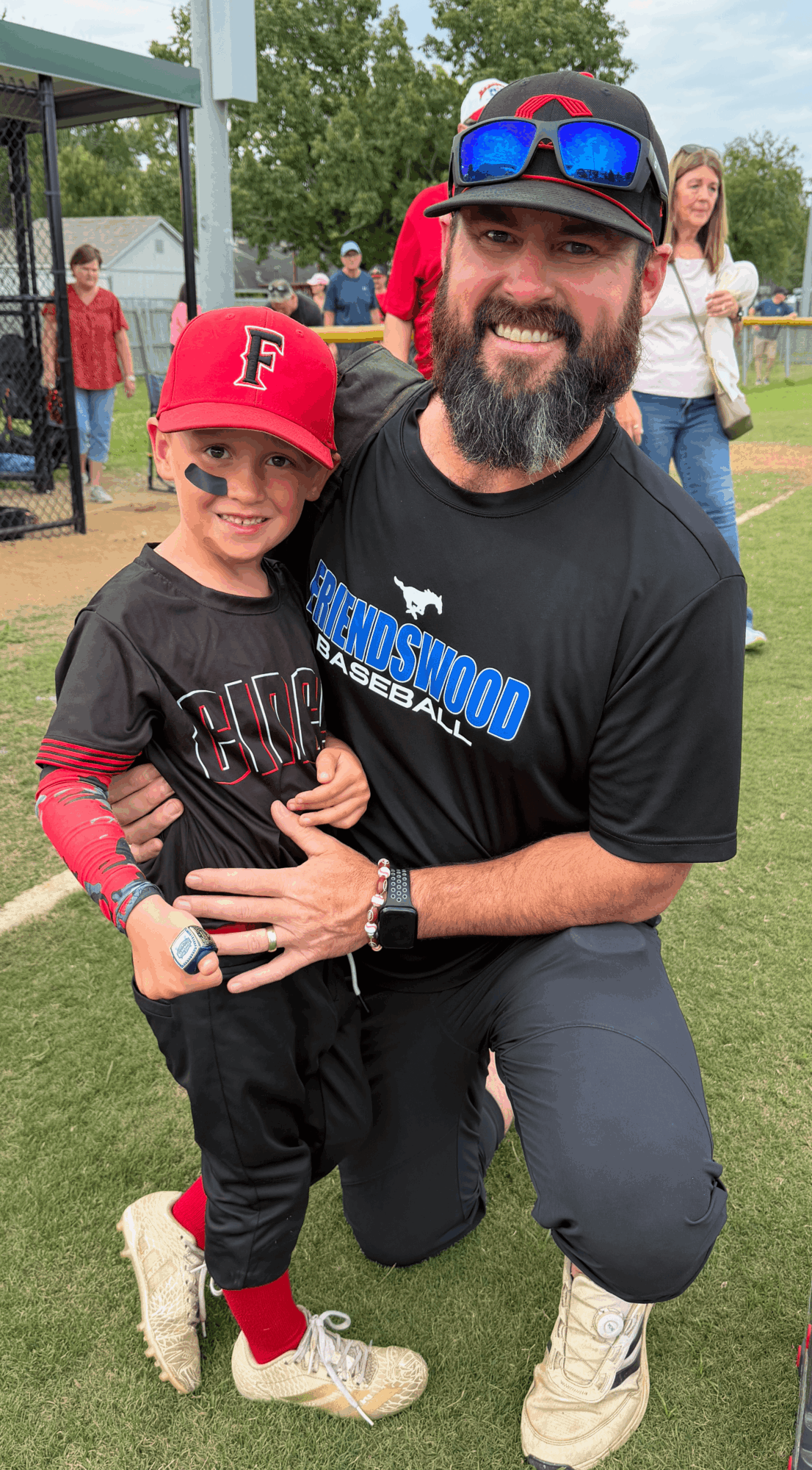 Man kneeling with a young boy outdoors, both smiling, wearing baseball caps and casual clothing, in a park setting.