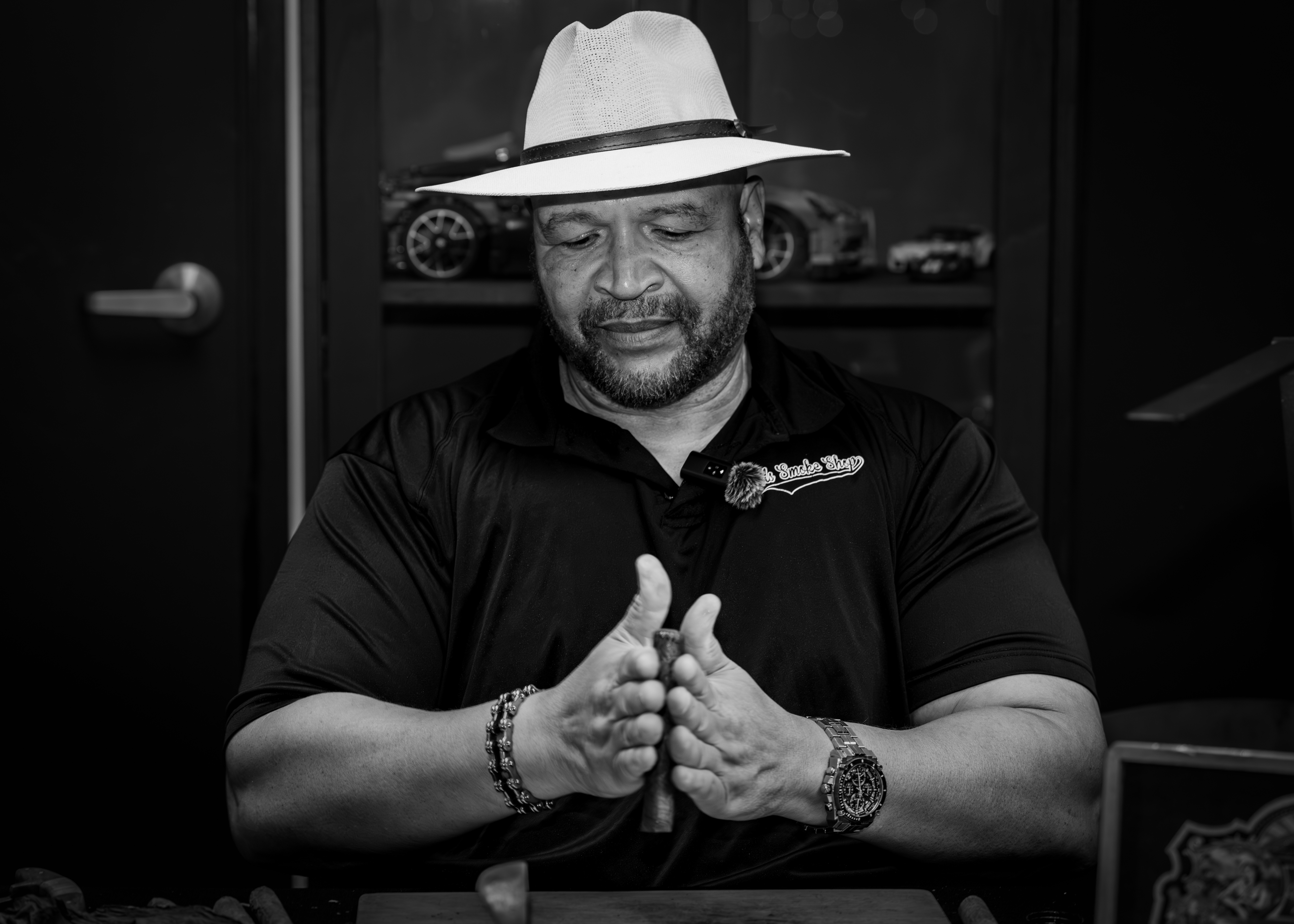 Man wearing a hat and dark shirt, sitting with hands clasped, in a room with shelves and cars in the background.
