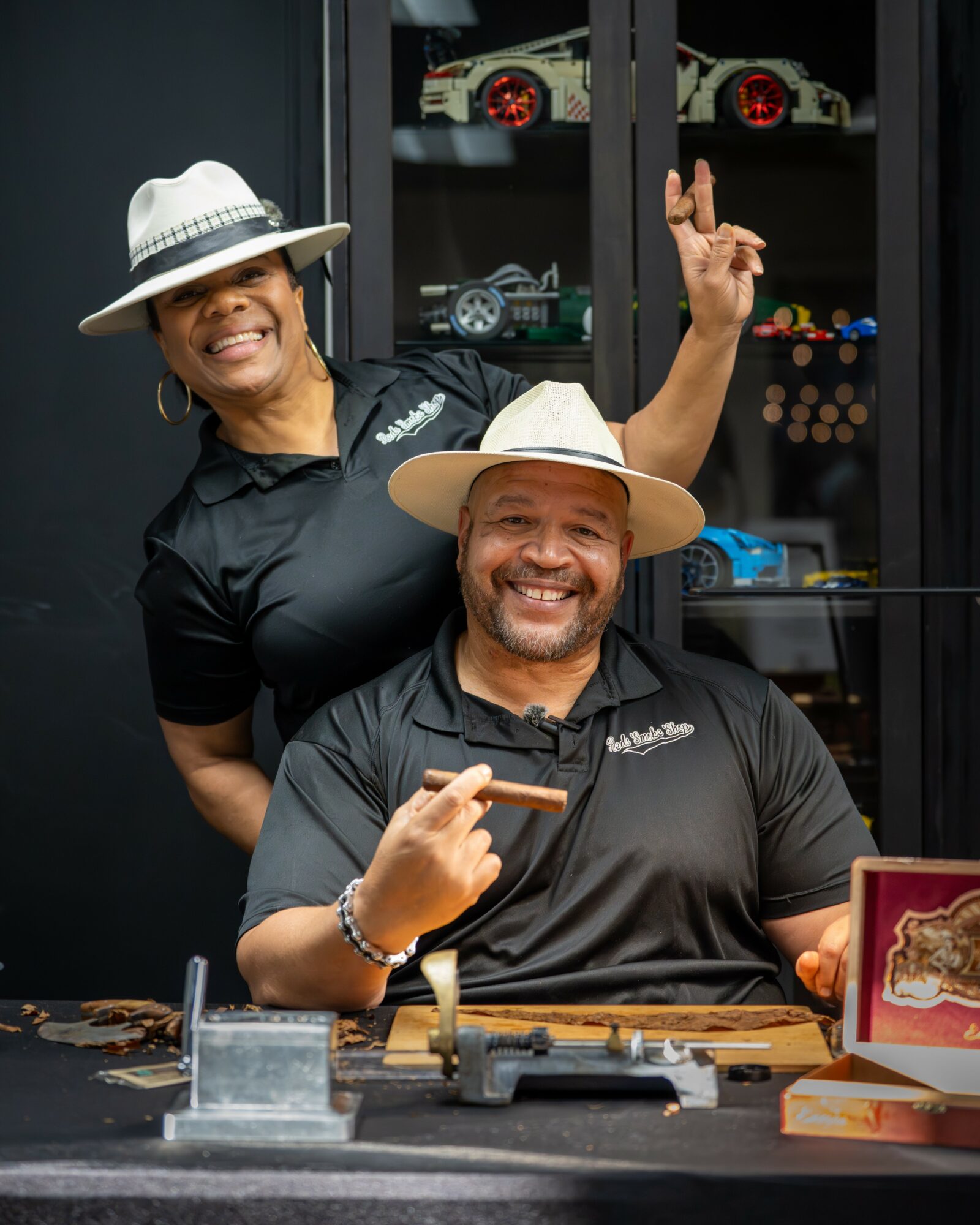 Two smiling people wearing hats, one seated and one standing, in front of a display case with model cars.