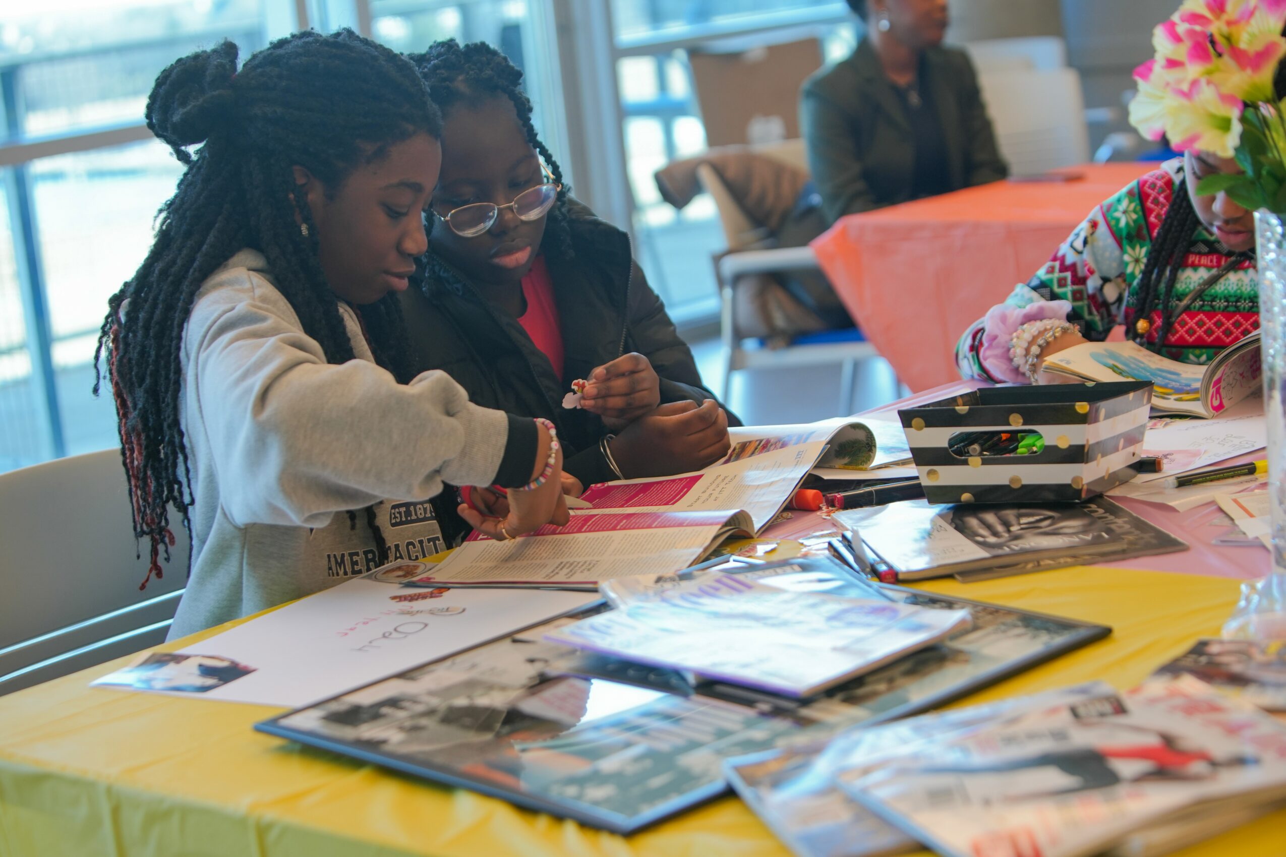 Two children sit at a table looking at books and papers, with a woman in the background. Bright room with large windows.