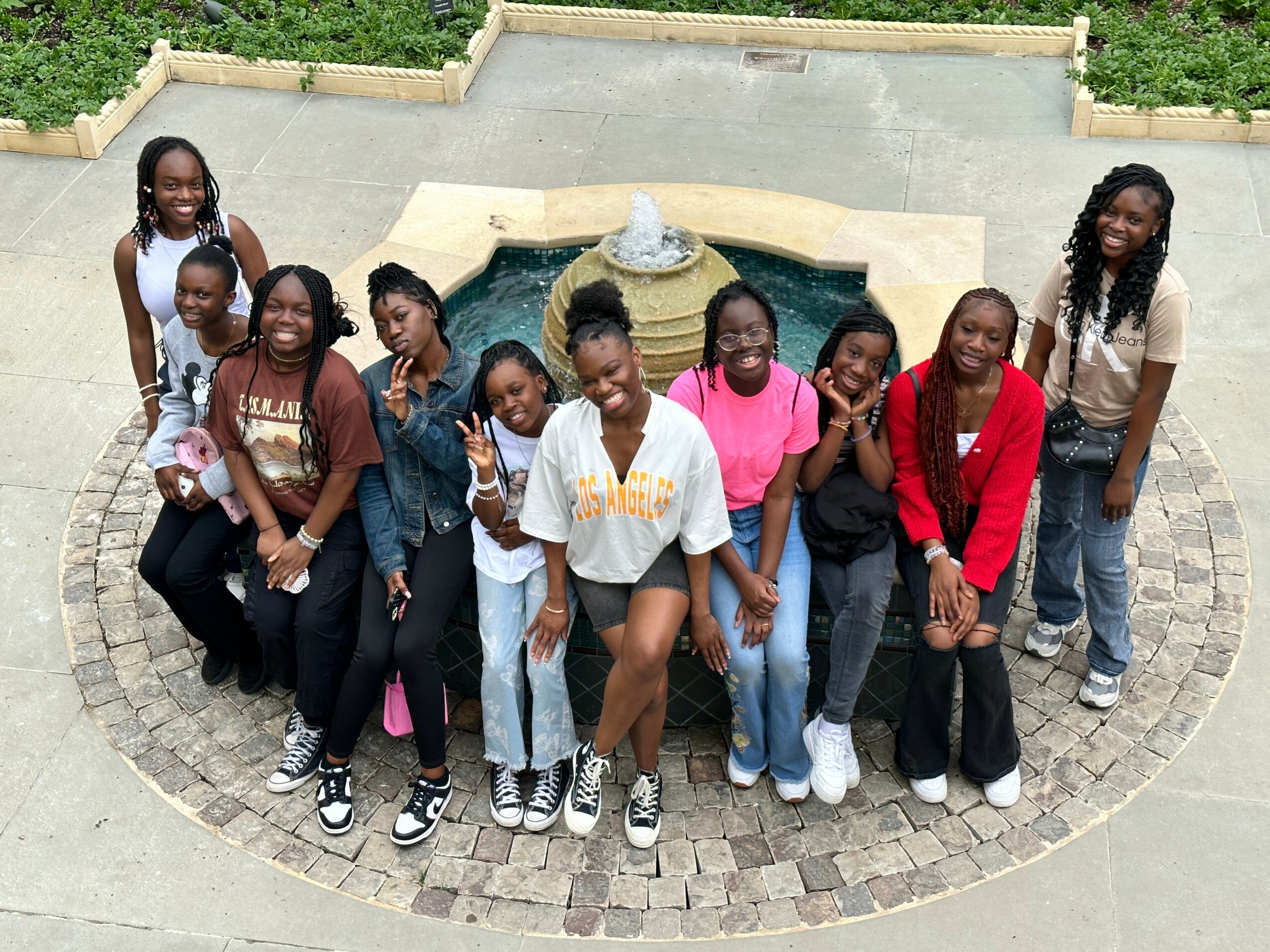 Group of ten children standing outdoors on a circular paved area with a fountain in the background.