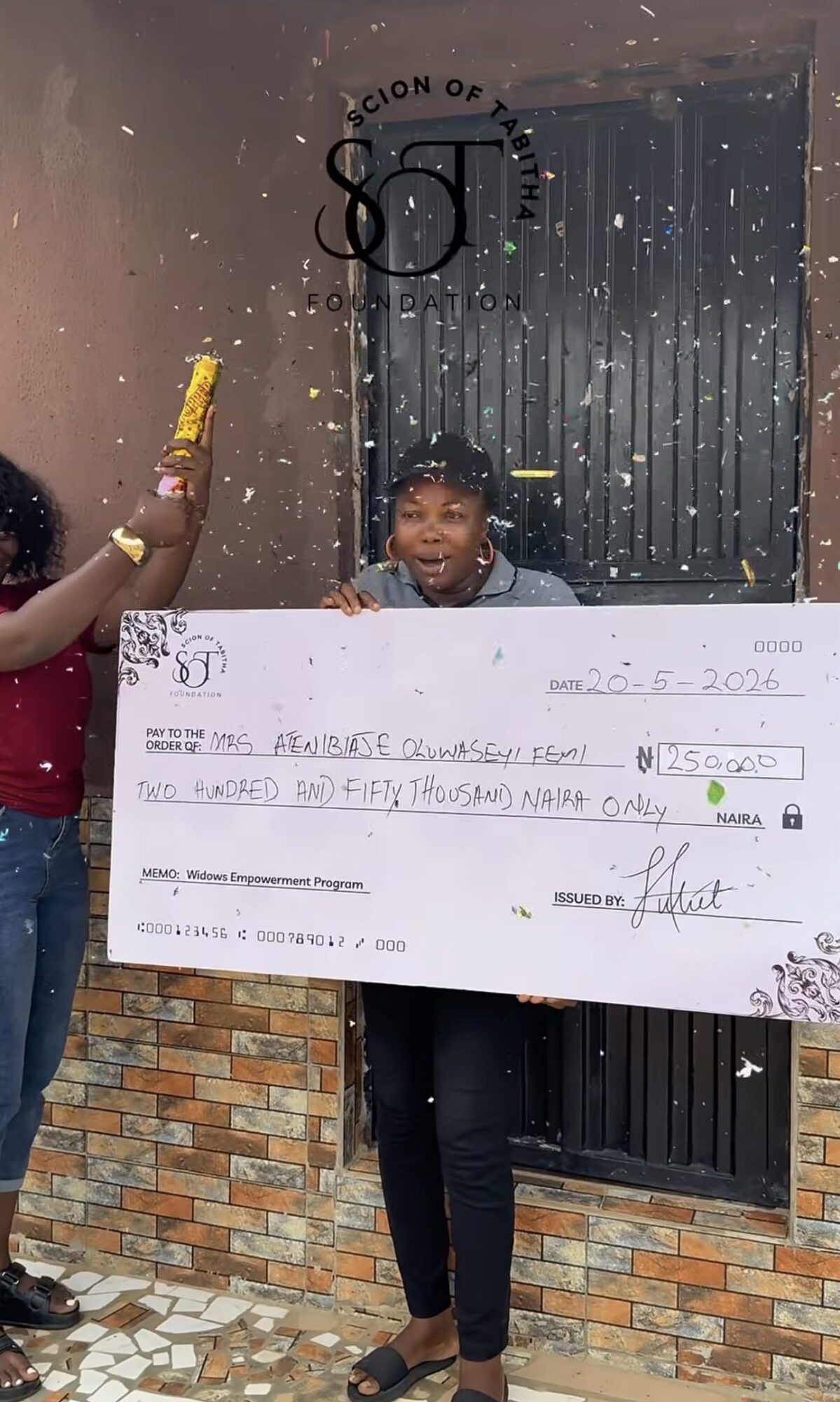 Child holding a large check with a person in the background, standing outside near a brick wall and black door.