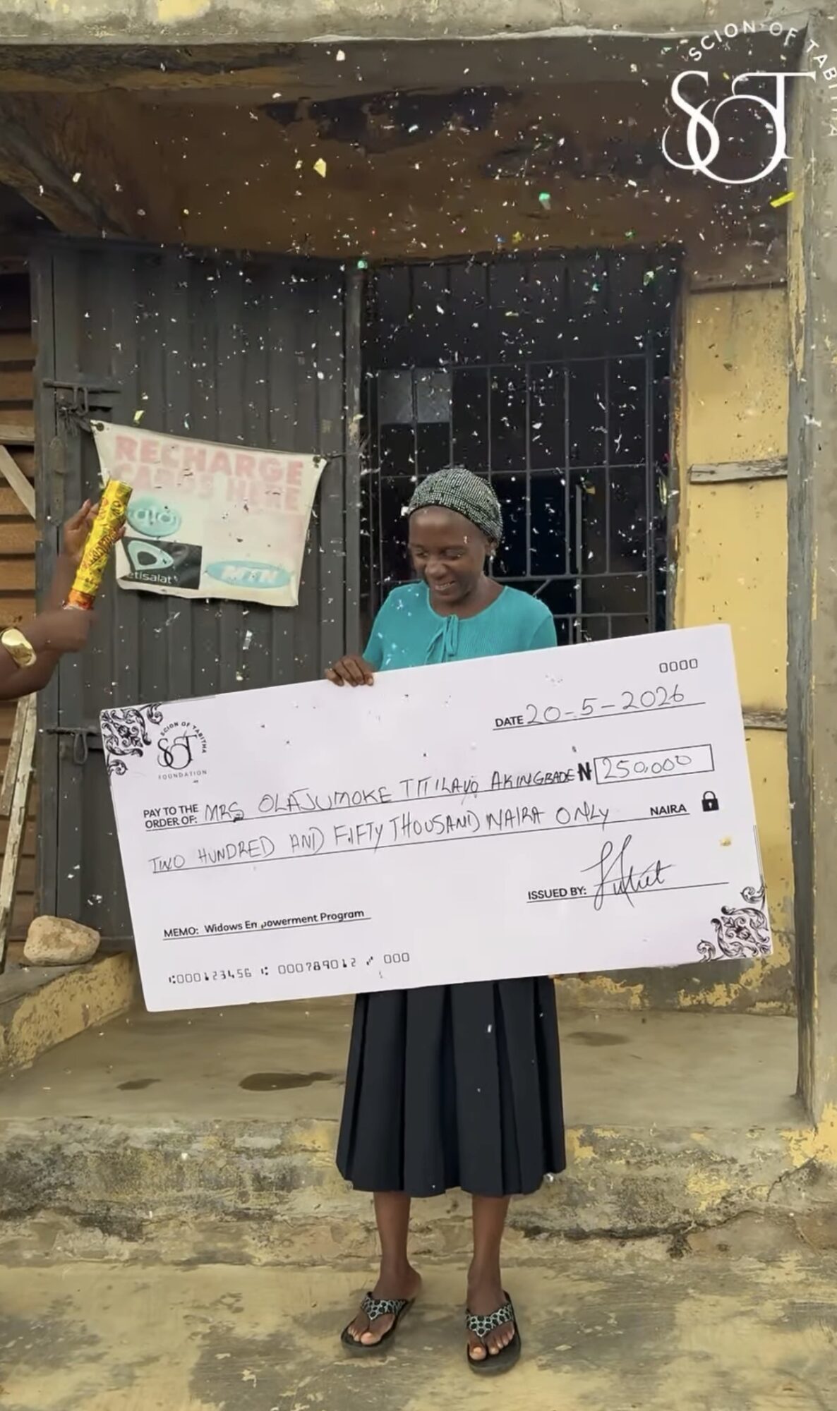 Woman holding a large check in front of a house, with a child and a sign in the background.