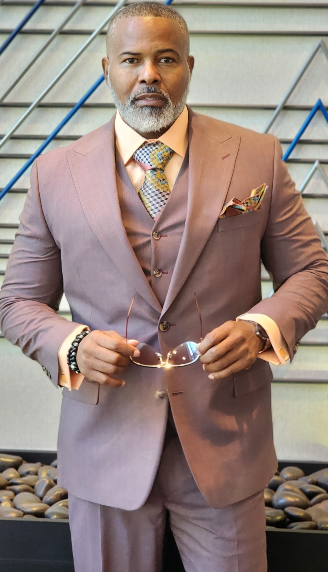 Man in a brown suit holding glasses, standing outdoors near stairs, with a serious expression.