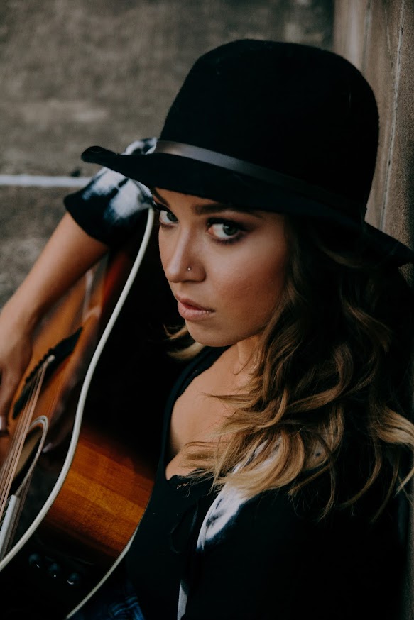 Woman wearing a black hat and black jacket, holding a guitar, looking at the camera, leaning against a wall.