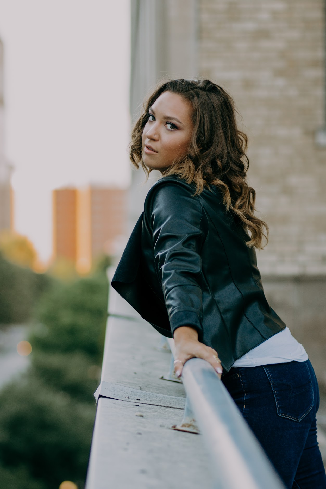 Woman with wavy hair leaning on a railing outdoors, city buildings in the background.