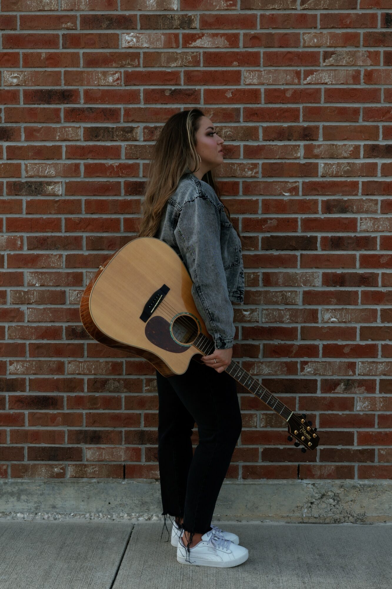 Young woman with long hair holding an acoustic guitar, standing sideways against a brick wall, wearing a denim jacket and white sneakers.