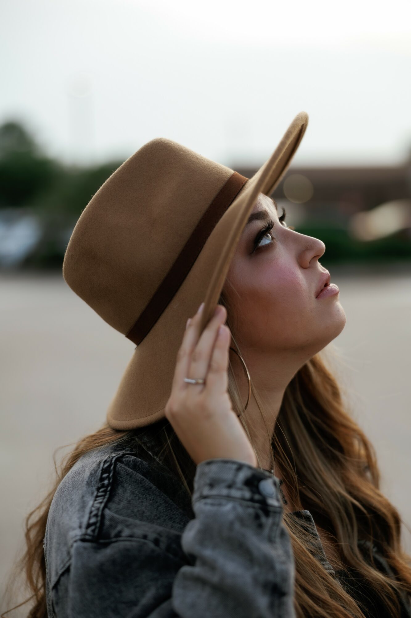 Woman wearing a wide-brimmed hat and denim jacket, looking upward, outdoors, blurred background.