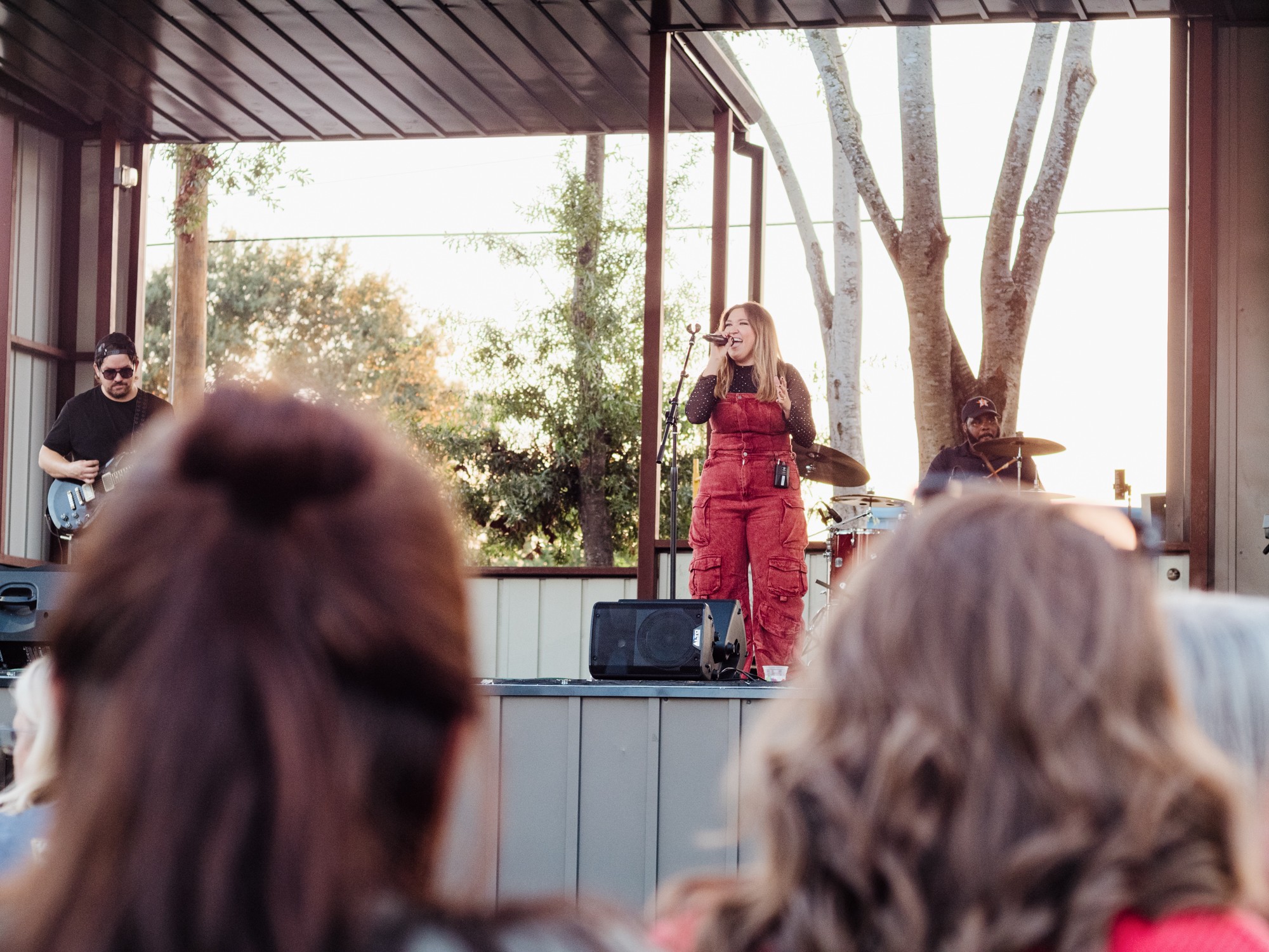 A woman in a red jumpsuit sings on an outdoor stage with trees and sunlight in the background, audience members in foreground.