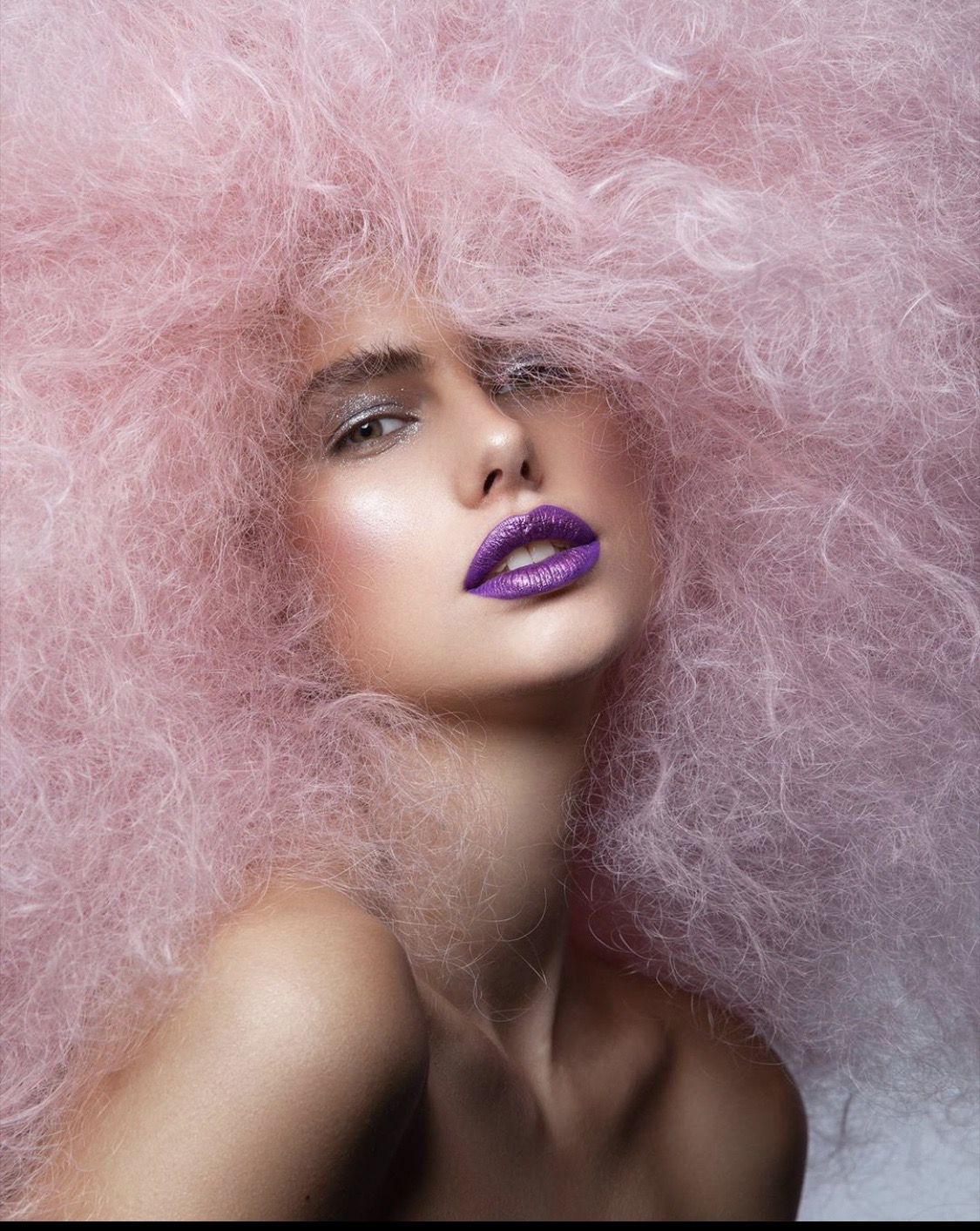 Woman with voluminous pink hair and purple lipstick, looking at the camera with a neutral expression.