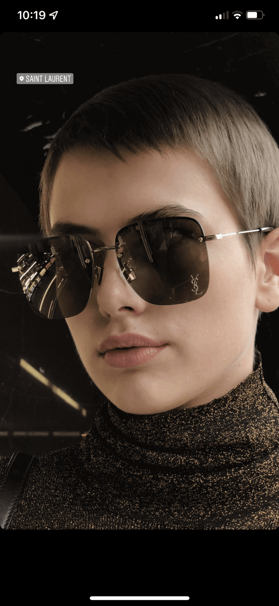 Young person with short hair wearing large sunglasses and a dark, glittery turtleneck, facing forward.